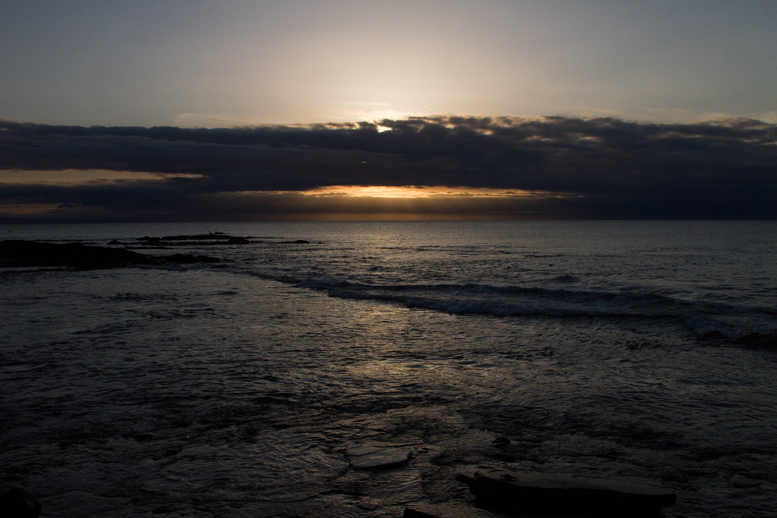 The sun setting behind low clouds over a rocky ocean shore, casting golden light across breaking waves.