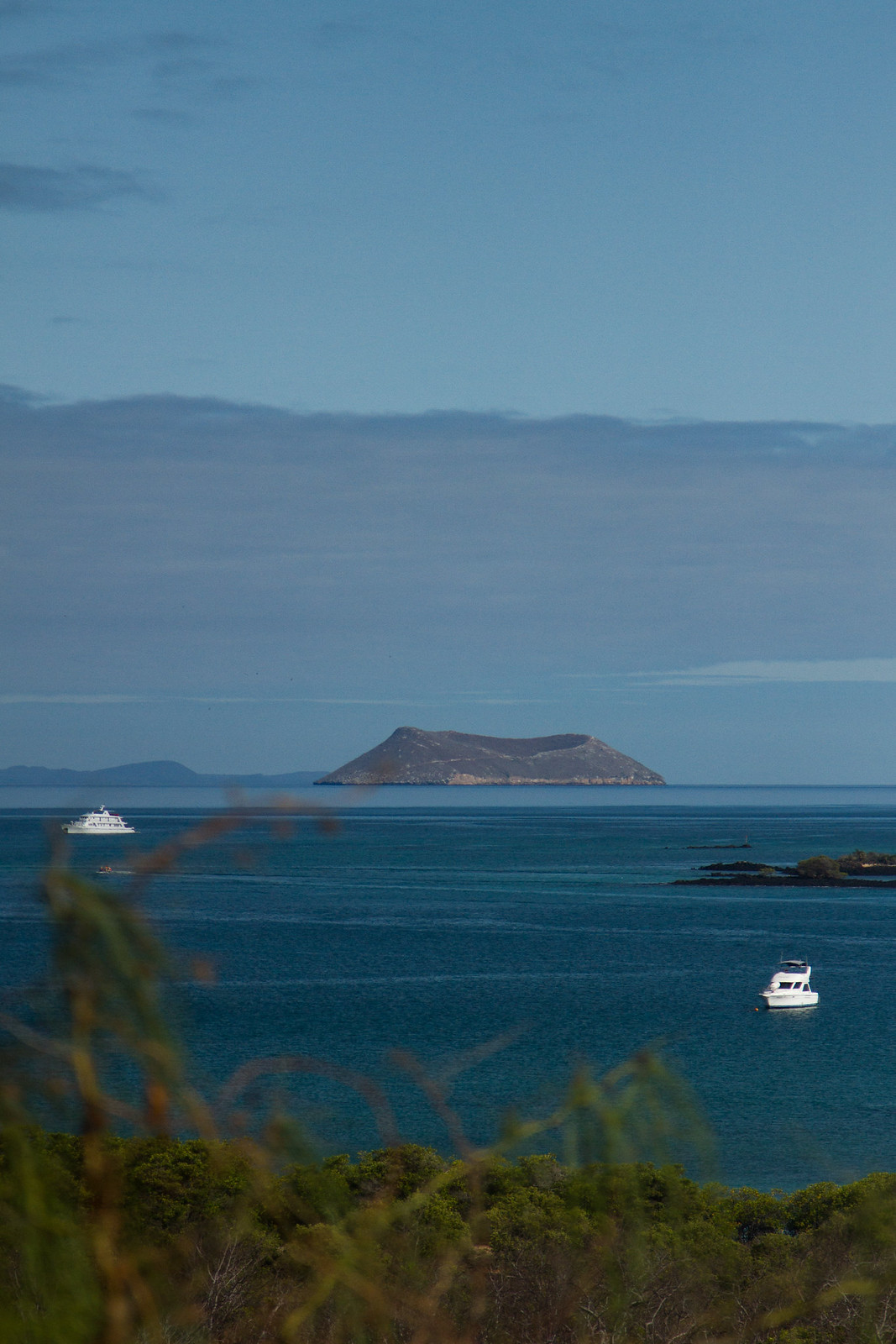 A small volcanic island on the horizon with two white yachts anchored in deep blue water, green brush in the foreground.