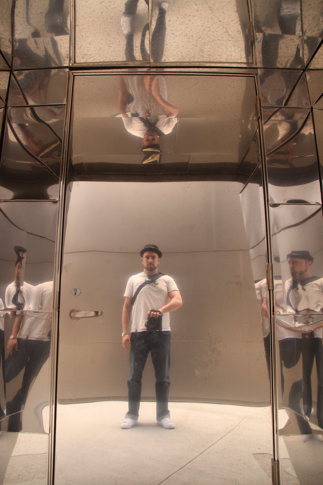 Man in a hat holding a camera, reflected many times in the mirrored steel walls of a doorway.