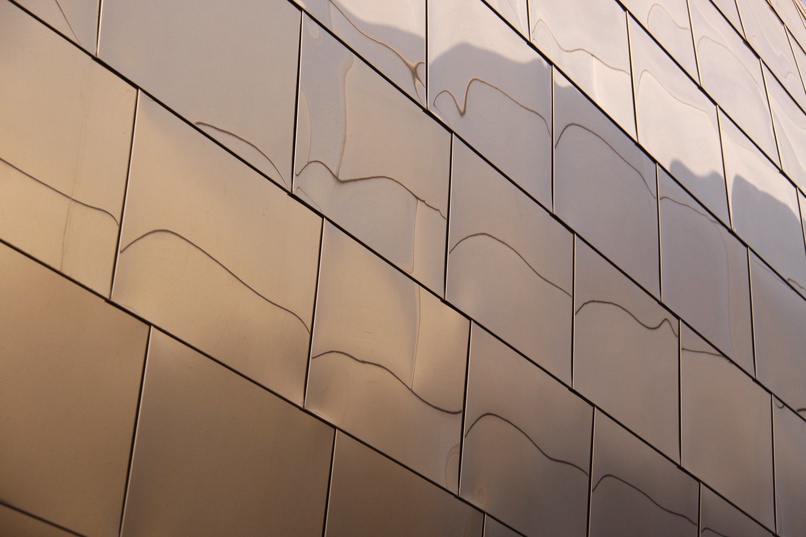 Sunlit stainless-steel panels of a Frank Gehry building, their rippled surfaces reflecting the sky in wavy diagonal lines.