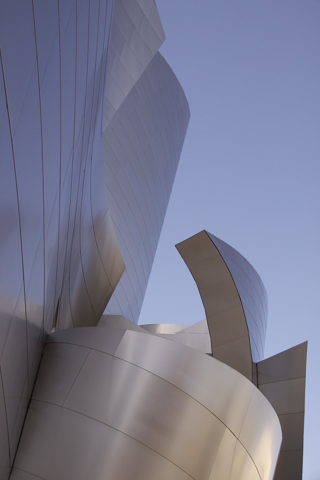 Curving stainless steel panels of a Frank Gehry building sweep upward against a clear blue sky.