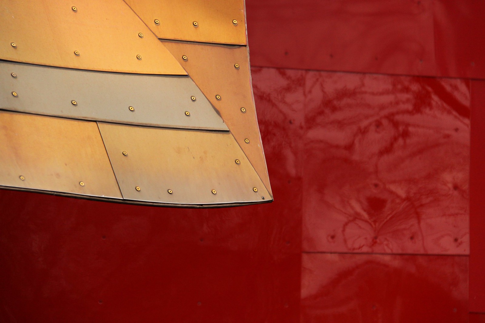 Curved, riveted metal panels in gold and steel set against a deep red glossy wall of Gehry's EMP building.