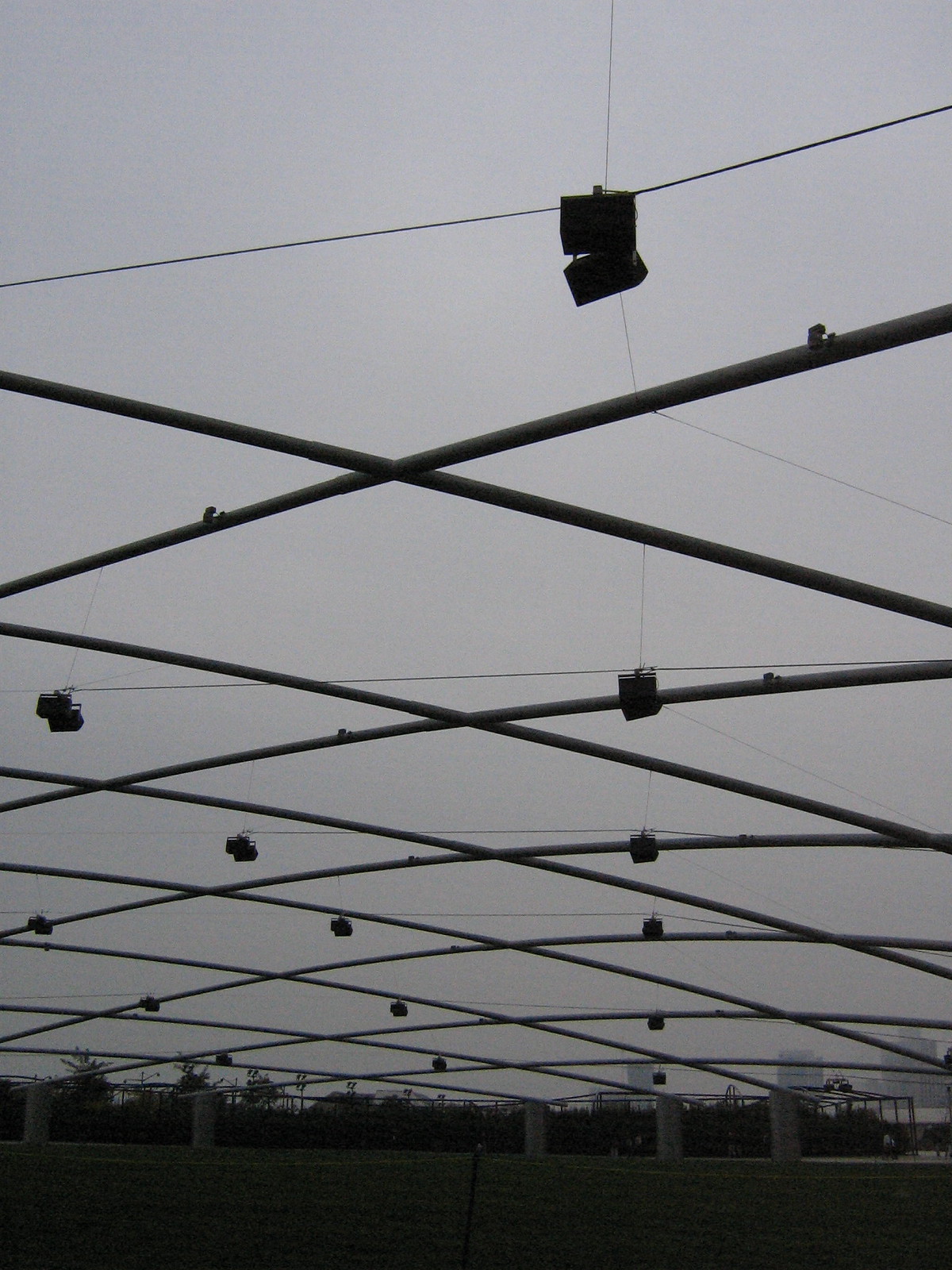 Criss-crossing steel trellis with hanging speakers over the Great Lawn of Millennium Park's Pritzker Pavilion under a grey sky.