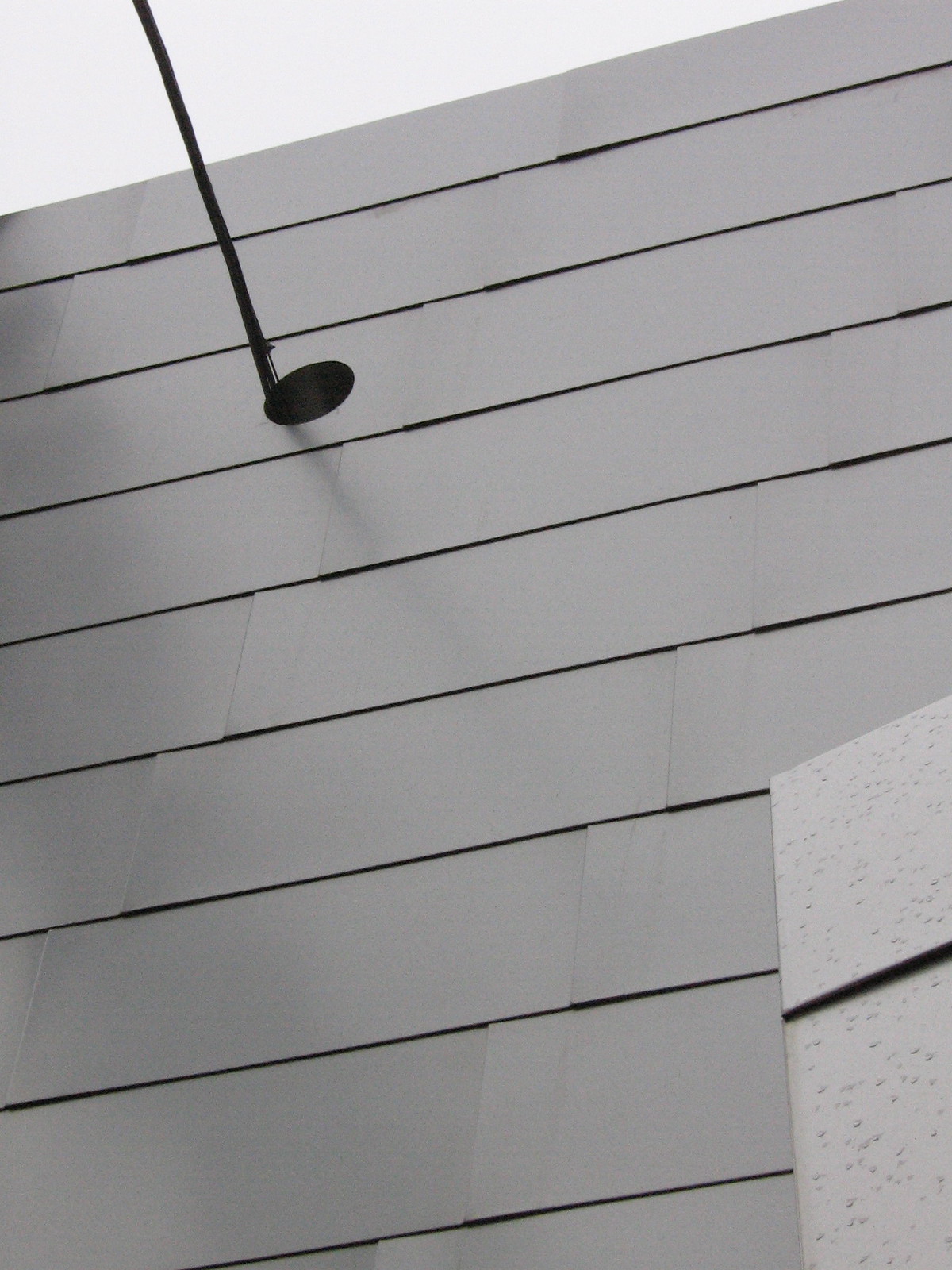 Overlapping grey metal panels clad a building facade against a pale sky, with a black light fixture jutting out.