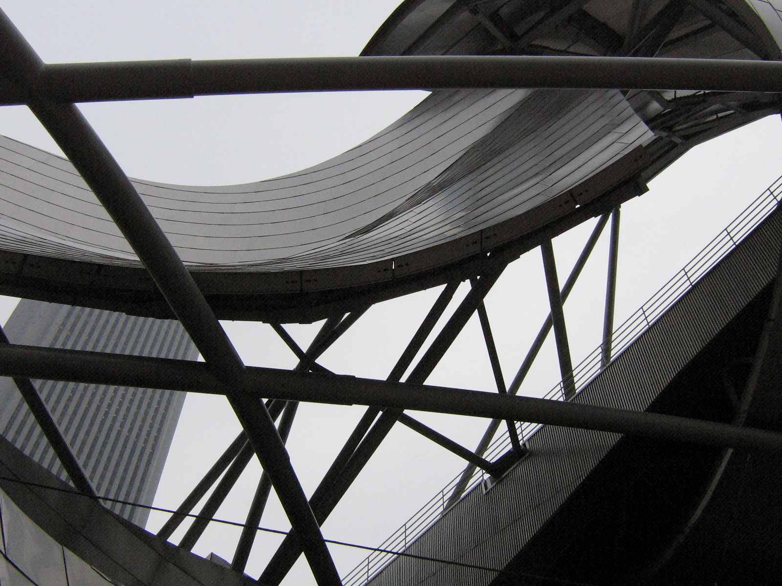 Criss-crossing steel beams support a curving metal canopy against a grey sky, viewed from below.