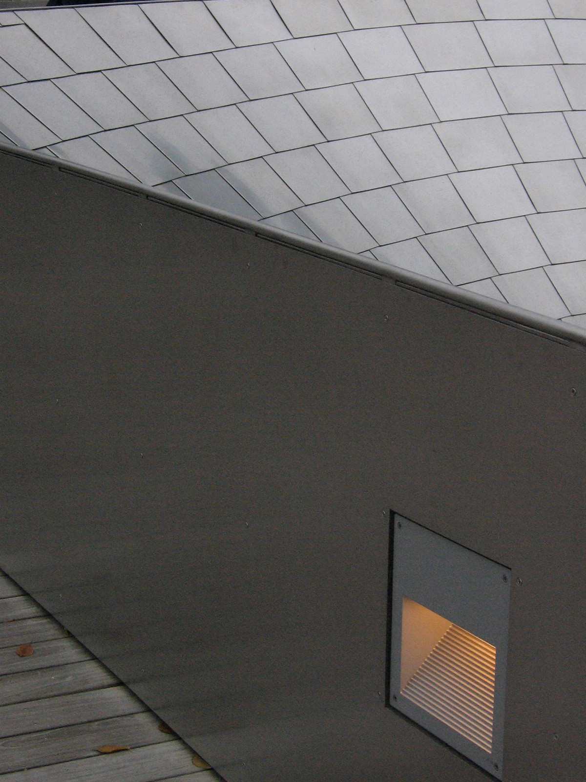 Curving metal-tiled roof meets a dark angular wall with a recessed amber step light above a wooden boardwalk.