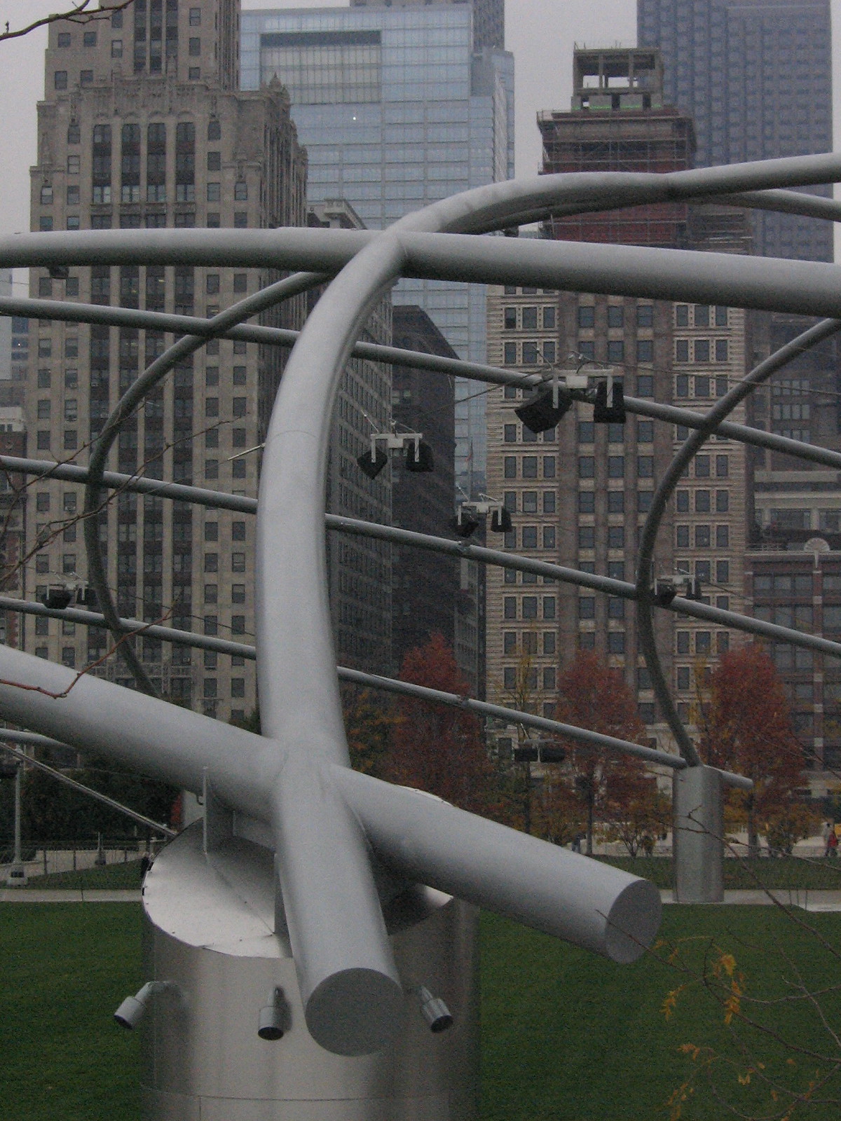Curved gray steel pipes of Frank Gehry's Pritzker Pavilion trellis arch over Millennium Park lawn against Chicago's skyline.