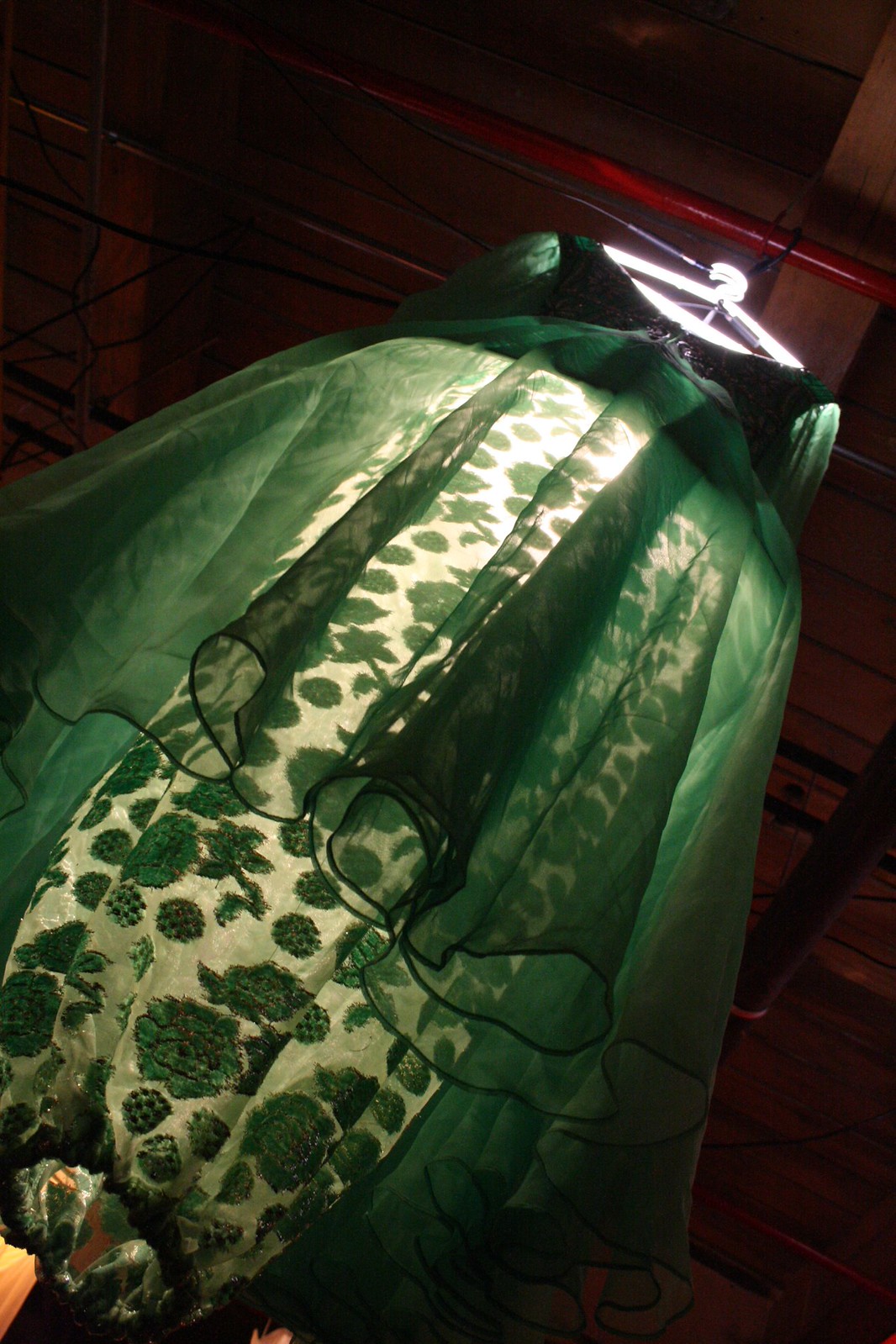 A green organza gown with velvet floral patterning hangs on a lit hanger beneath a wooden rafter ceiling.