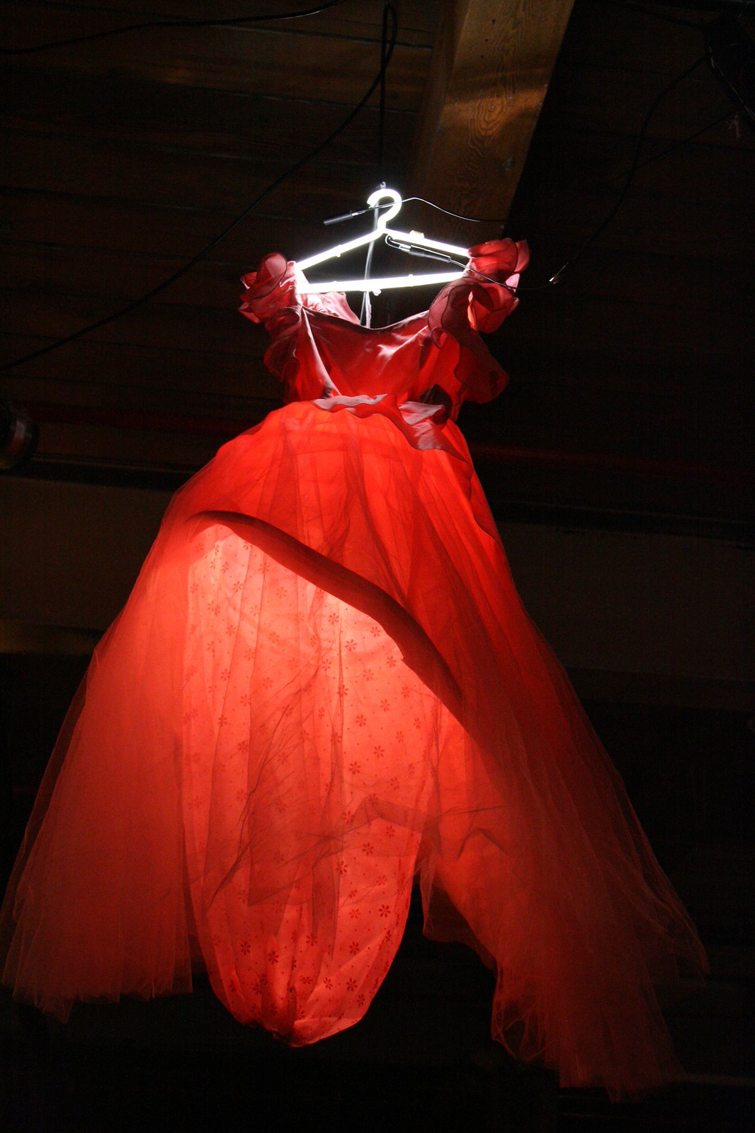 A glowing red tulle ball gown on a hanger lit from within by a fluorescent tube, in a dark rafters space.