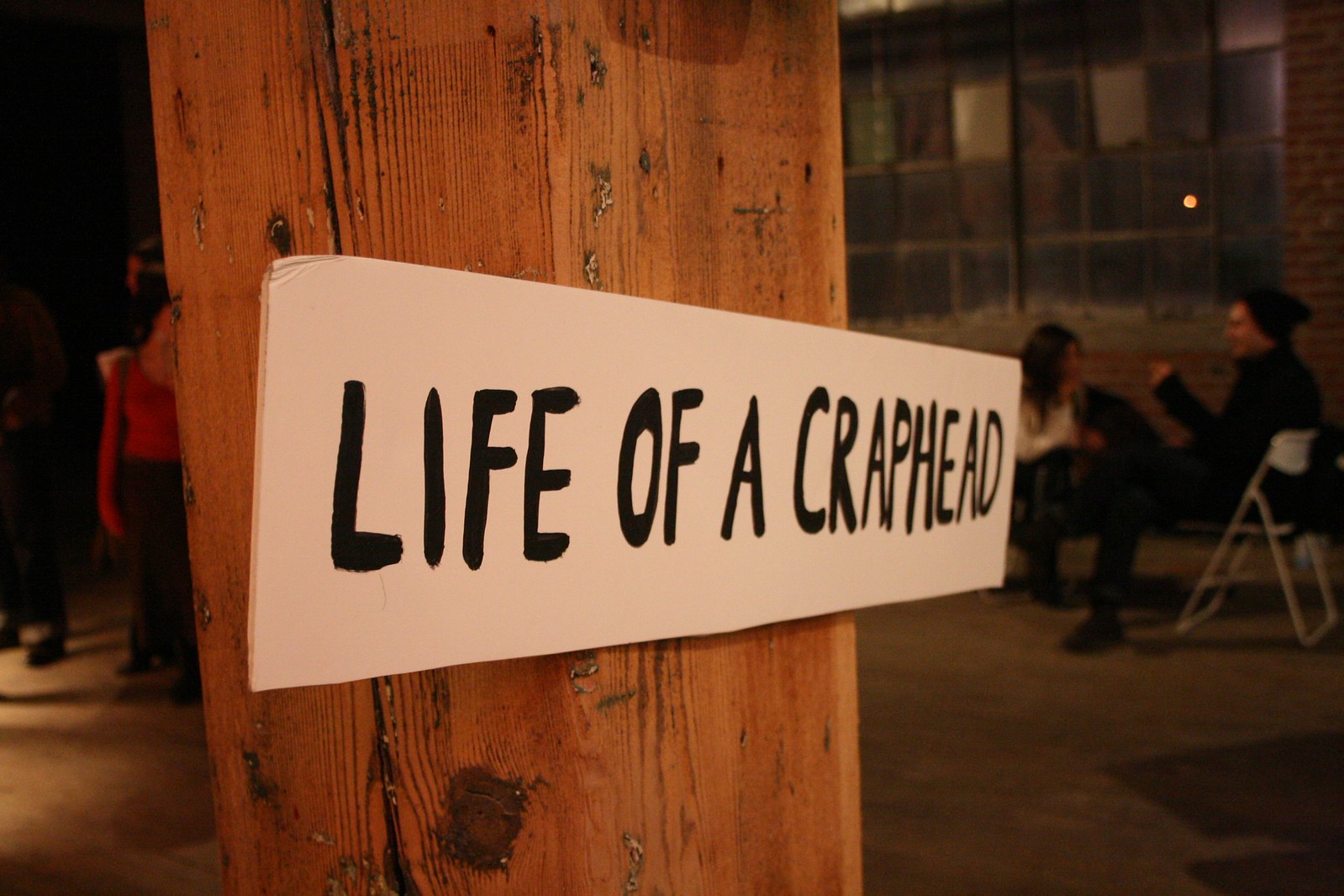 Hand-painted "LIFE OF A CRAPHEAD" sign on a wooden post in a dim warehouse with people seated behind.