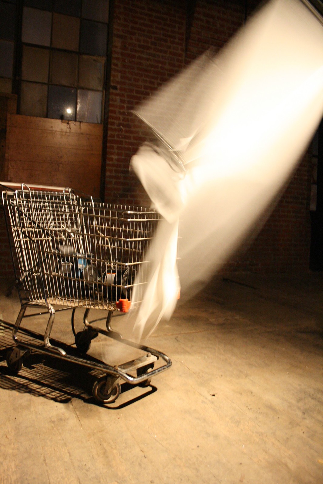 A shopping cart in a brick warehouse with a blurred sheet of white fabric flung across it in motion.