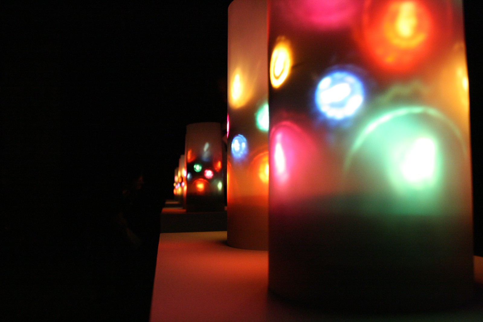 A row of frosted cylindrical lamps glowing with blurred spots of red, blue, green, and orange light in a dark room.