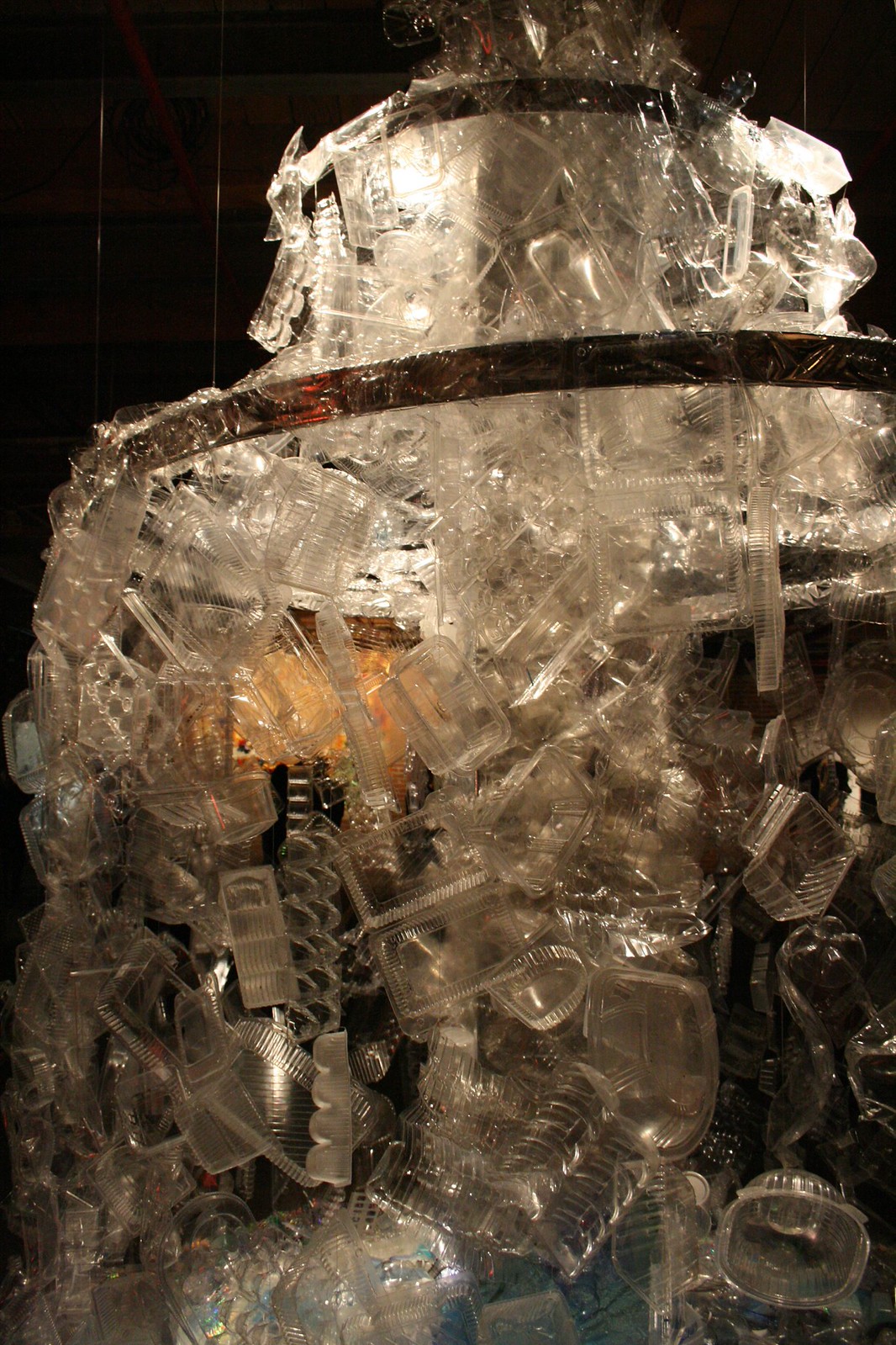 Towering chandelier-like sculpture built from clear plastic clamshell containers and packaging, lit from within.