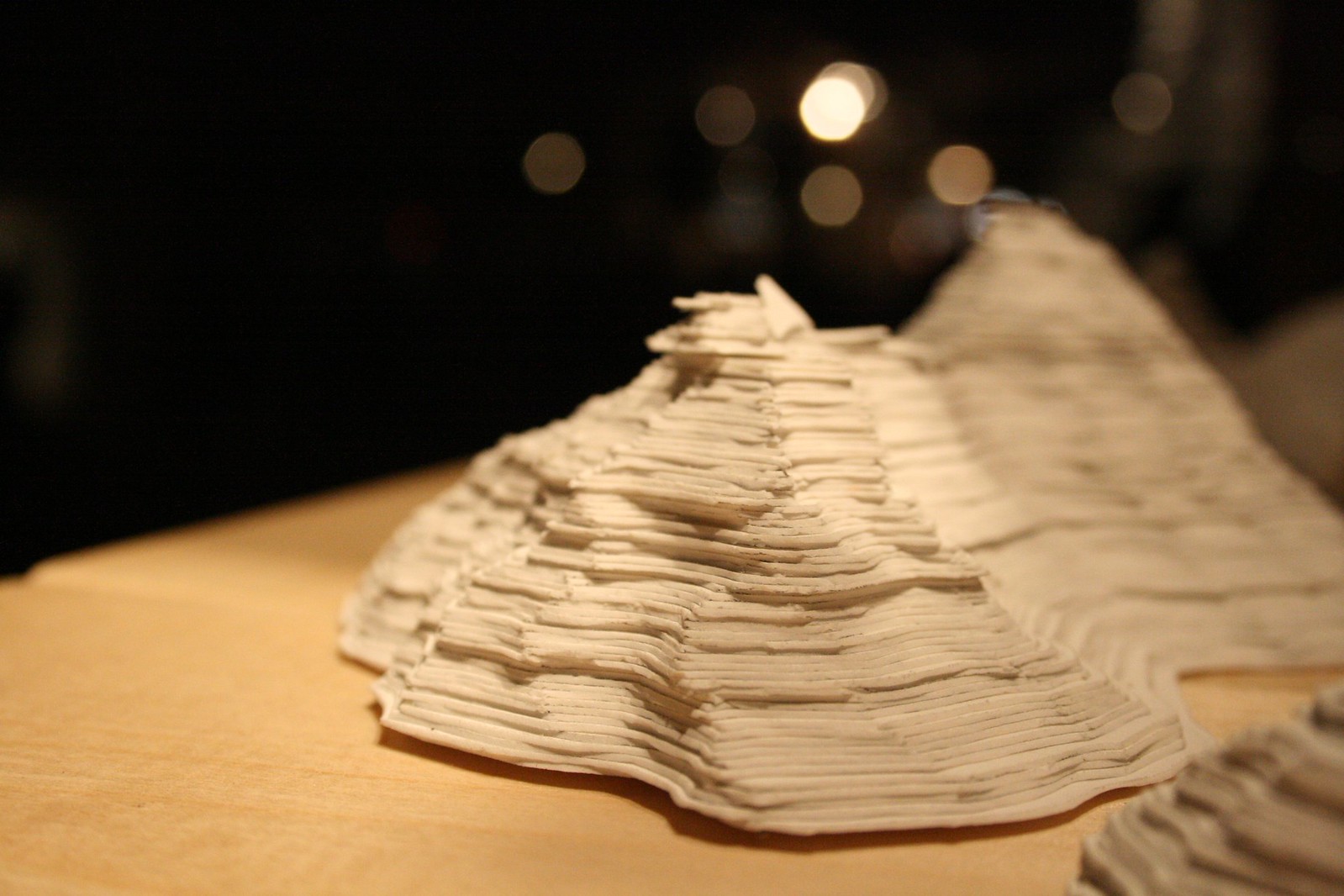 Layered contour relief model of a mountain built from stacked paper sheets, lit against a dark background.