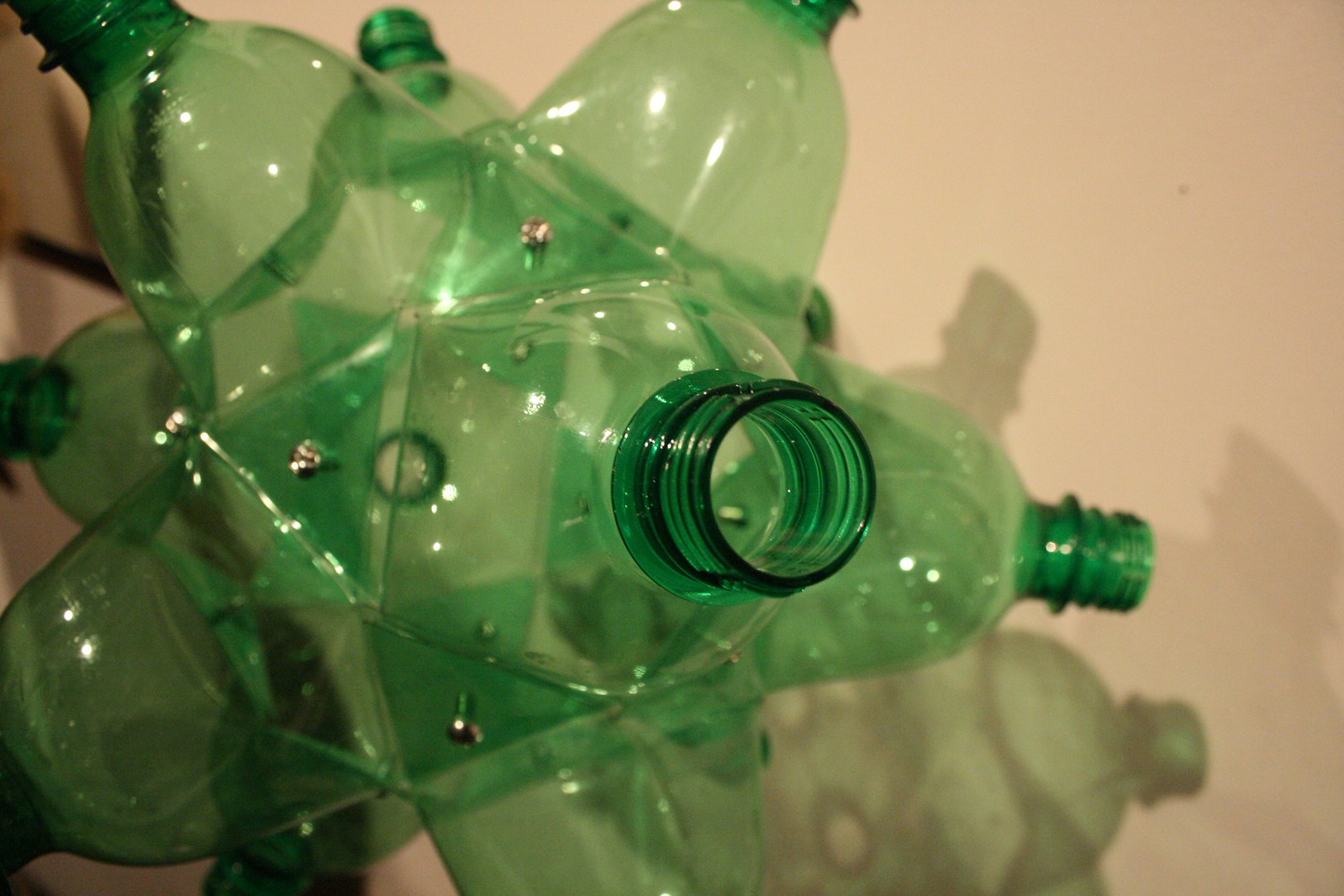 Sculpture of green plastic bottles bolted together into a spiky cluster, casting shadows on a pale wall.