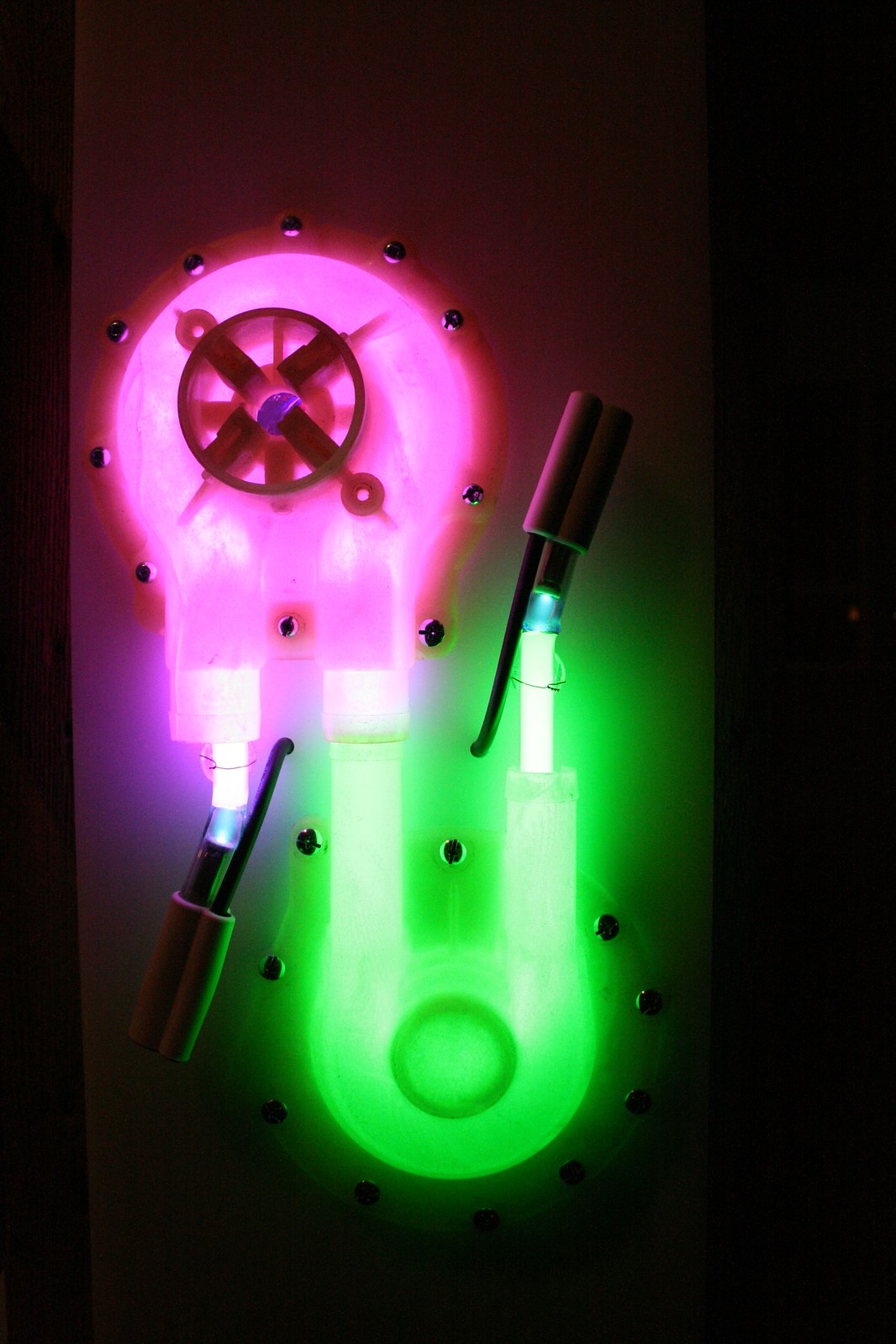 Wall-mounted sculpture of translucent pump housings glowing pink and green with neon tubes in a darkened room.