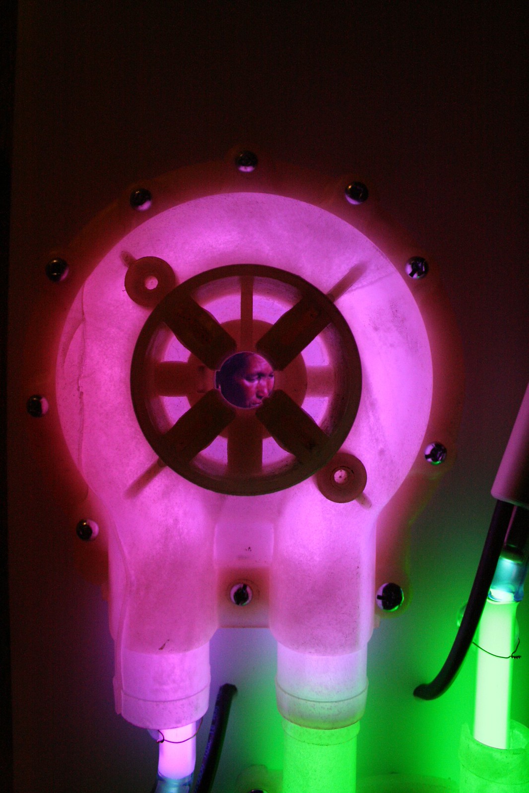 A person's face reflected in the center of a glowing pink water pump beside a green neon tube.