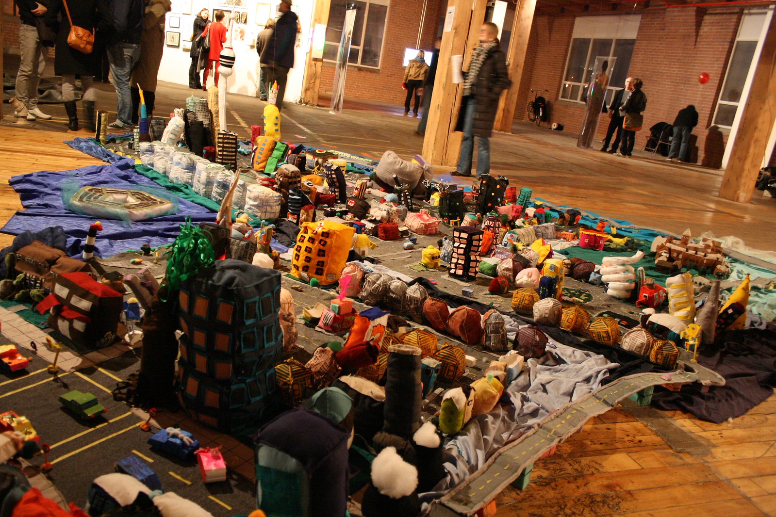 A sprawling fabric miniature city of soft sculpted buildings on a gallery floor as visitors mingle.