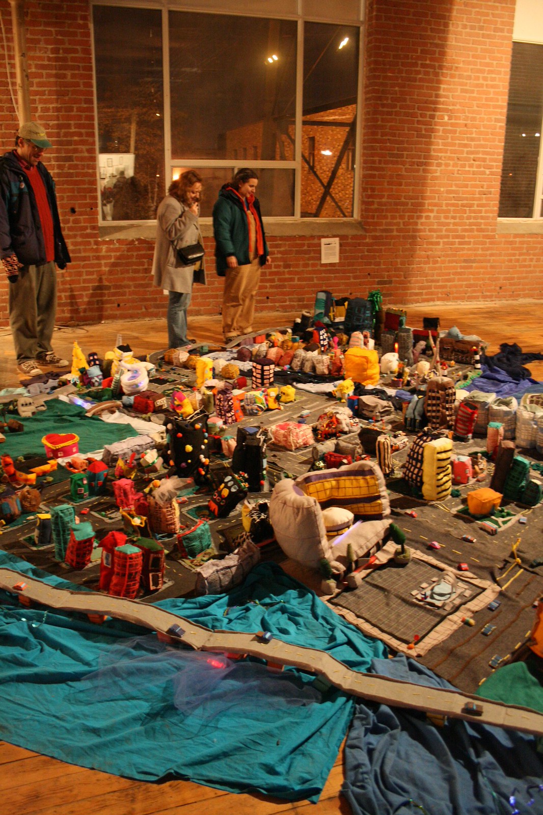 Gallery visitors examine a sprawling floor installation of a soft-sculpture city, buildings sewn from fabric, in a brick loft.