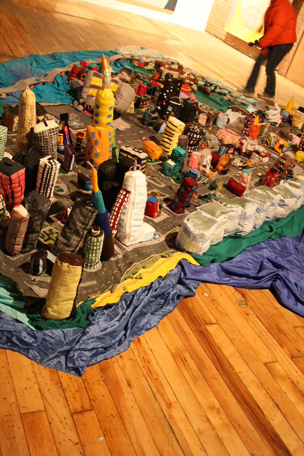 A soft-sculpture fabric cityscape of stuffed skyscrapers on a gallery floor, with a visitor walking past.