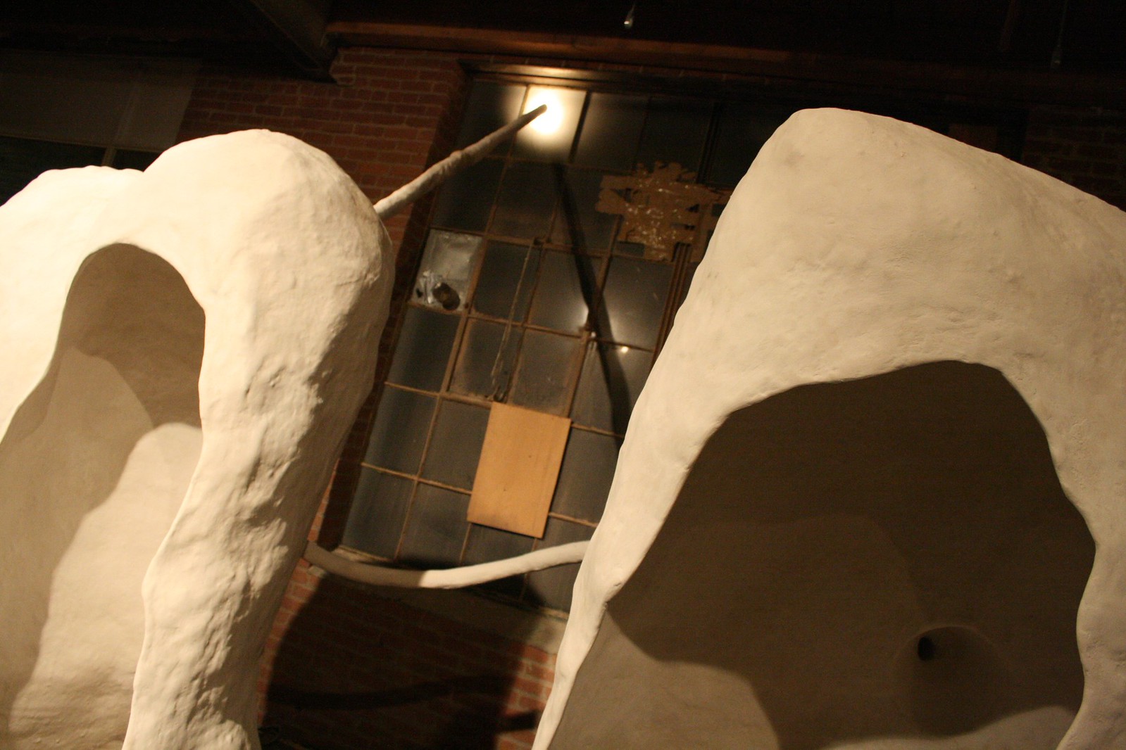 Two large white organic plaster sculptures flank a dim brick gallery wall with a lit window.