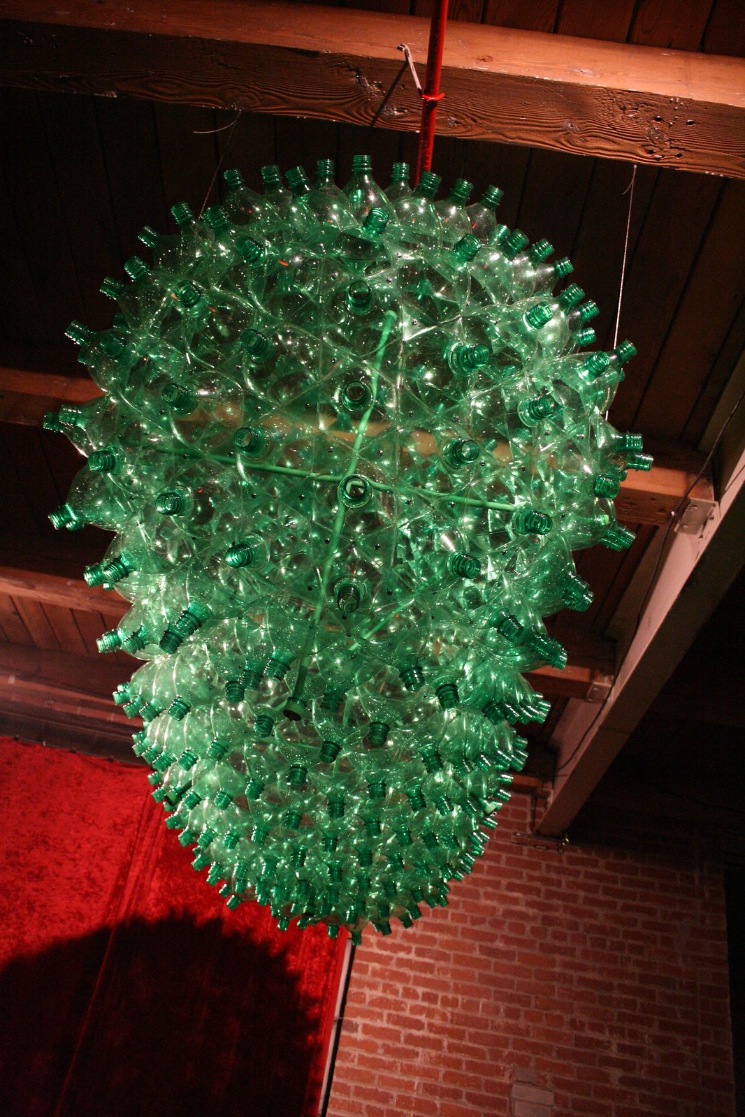 Suspended sculpture of clustered green plastic bottles hanging from wooden ceiling beams above a brick wall.