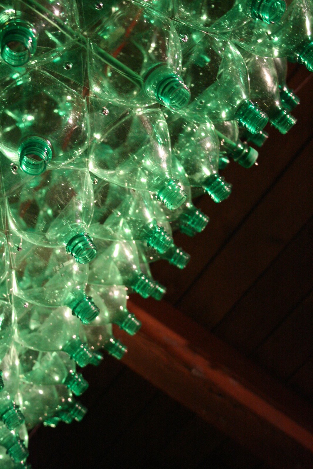 Backlit sculpture of clustered green plastic bottles glowing against a dark wooden ceiling.