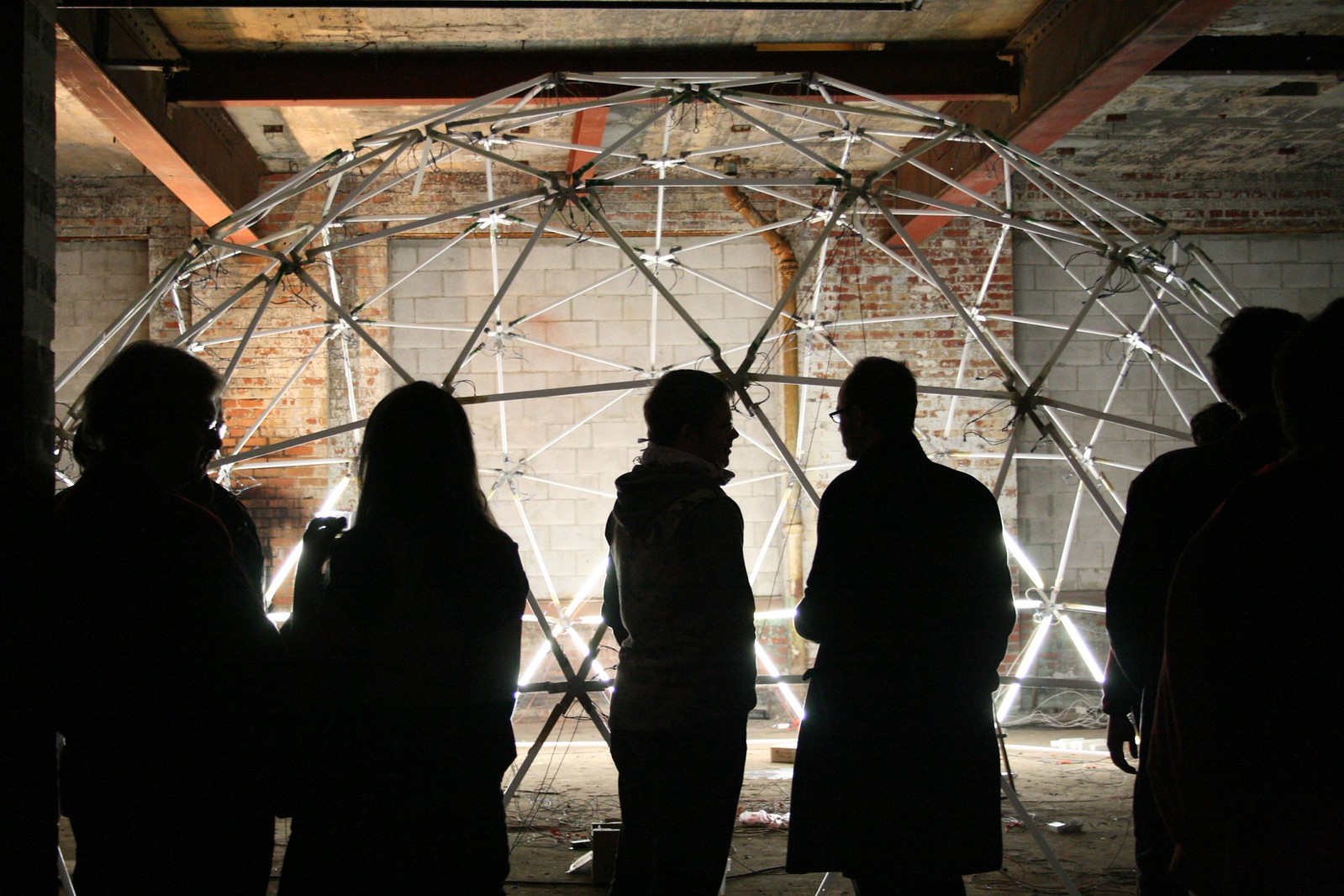 Silhouetted people gather before a glowing geodesic dome sculpture in a raw brick warehouse space.