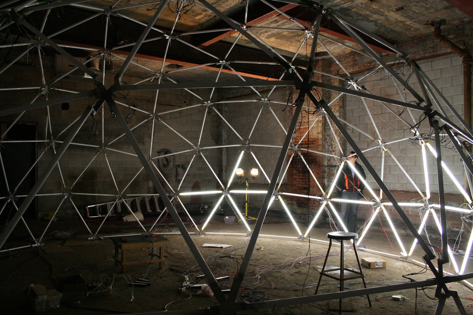 A person builds a geodesic dome laced with fluorescent tubes inside a dim, gutted brick warehouse.