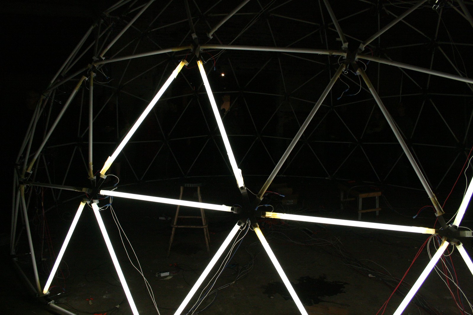 A geodesic dome sculpture lit by glowing fluorescent tubes in a dark warehouse space.