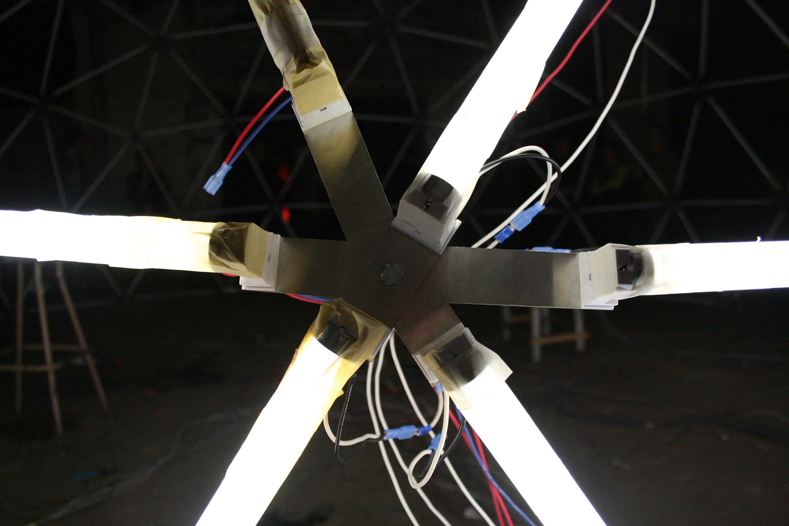 Six glowing fluorescent tubes radiate from a metal hub with tangled wires, inside a dark geodesic dome.