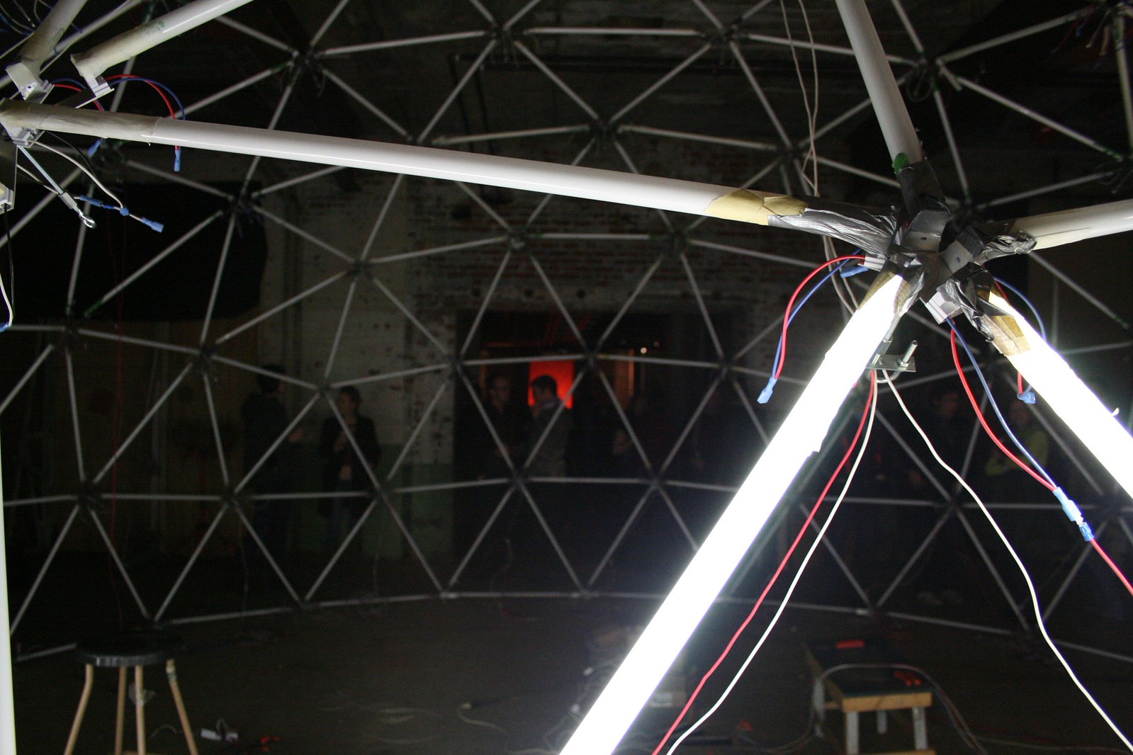 Geodesic dome of white poles and colored wiring lit by fluorescent tubes, with people gathered in a dim brick warehouse beyond.