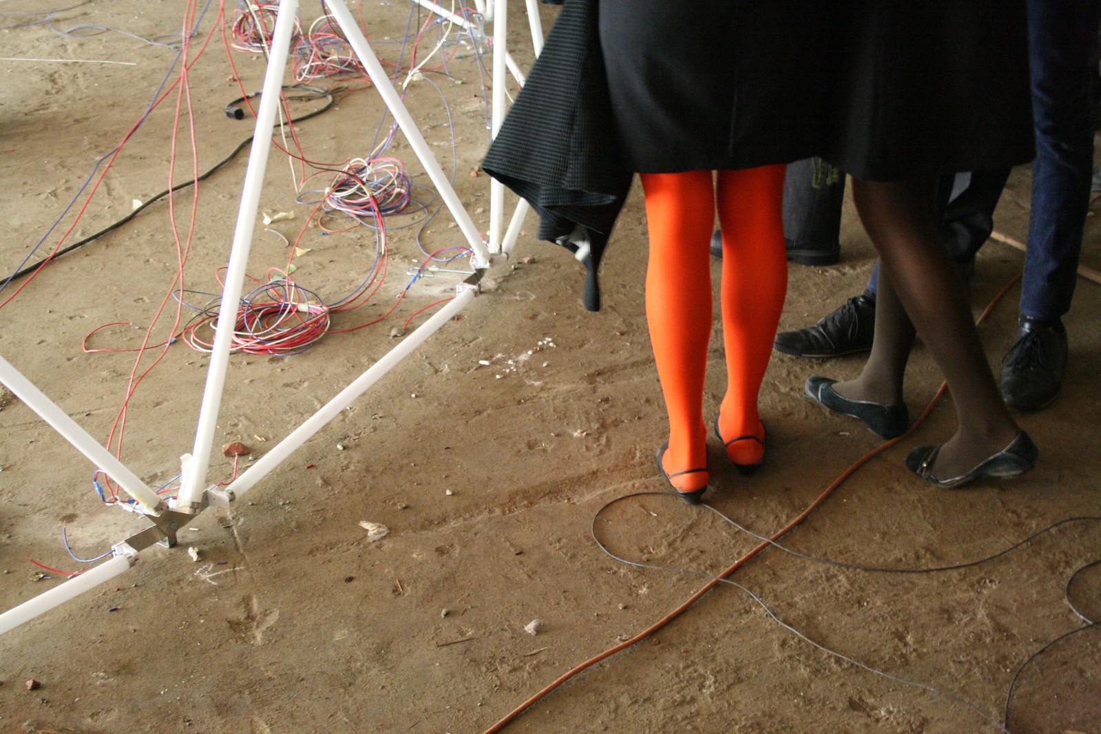 People's legs beside a white pyramid sculpture and tangled colored wires on a dirt floor, one in orange tights.