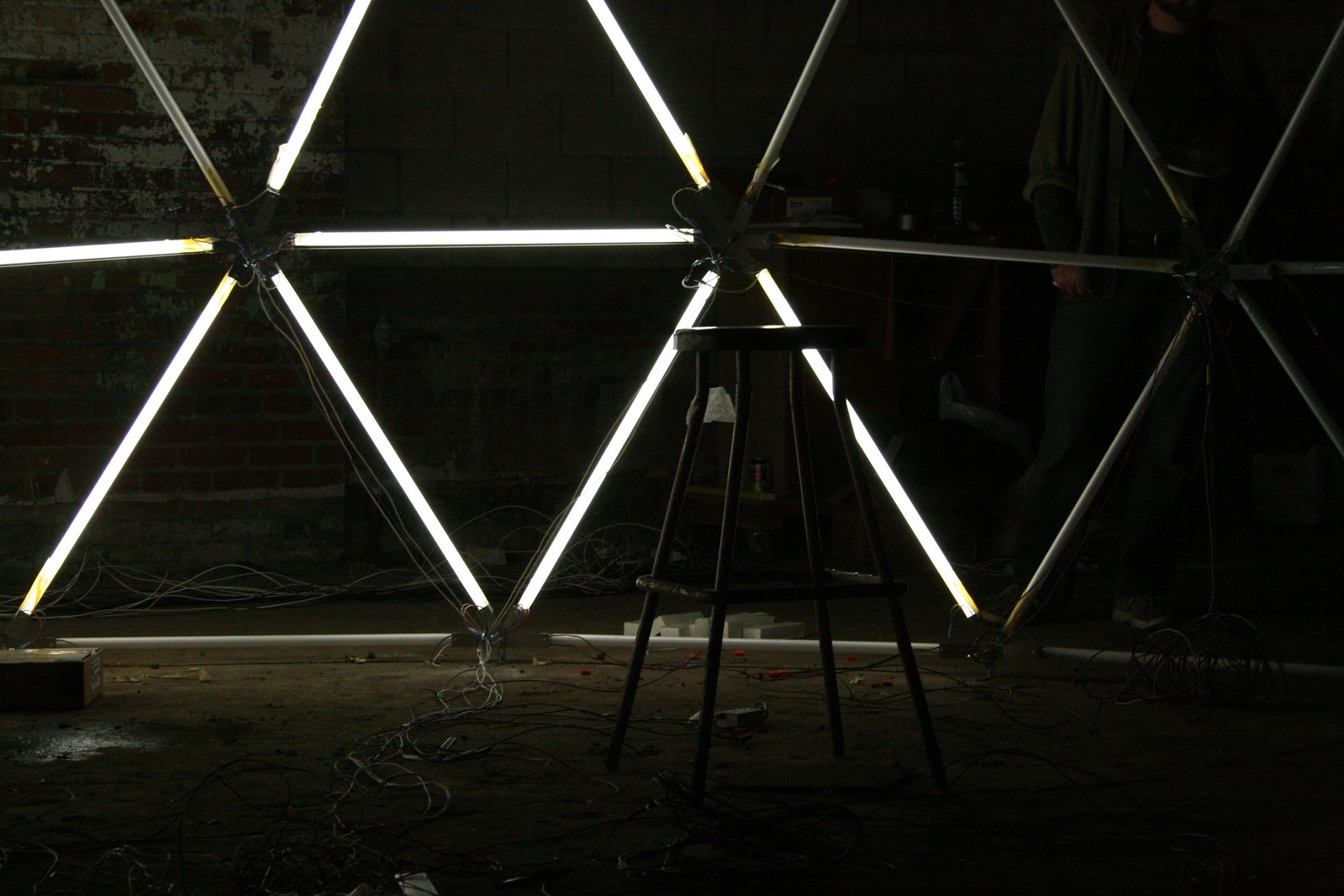 Wooden stool before a triangular fluorescent-tube light sculpture with tangled wires in a dim brick-walled workshop.