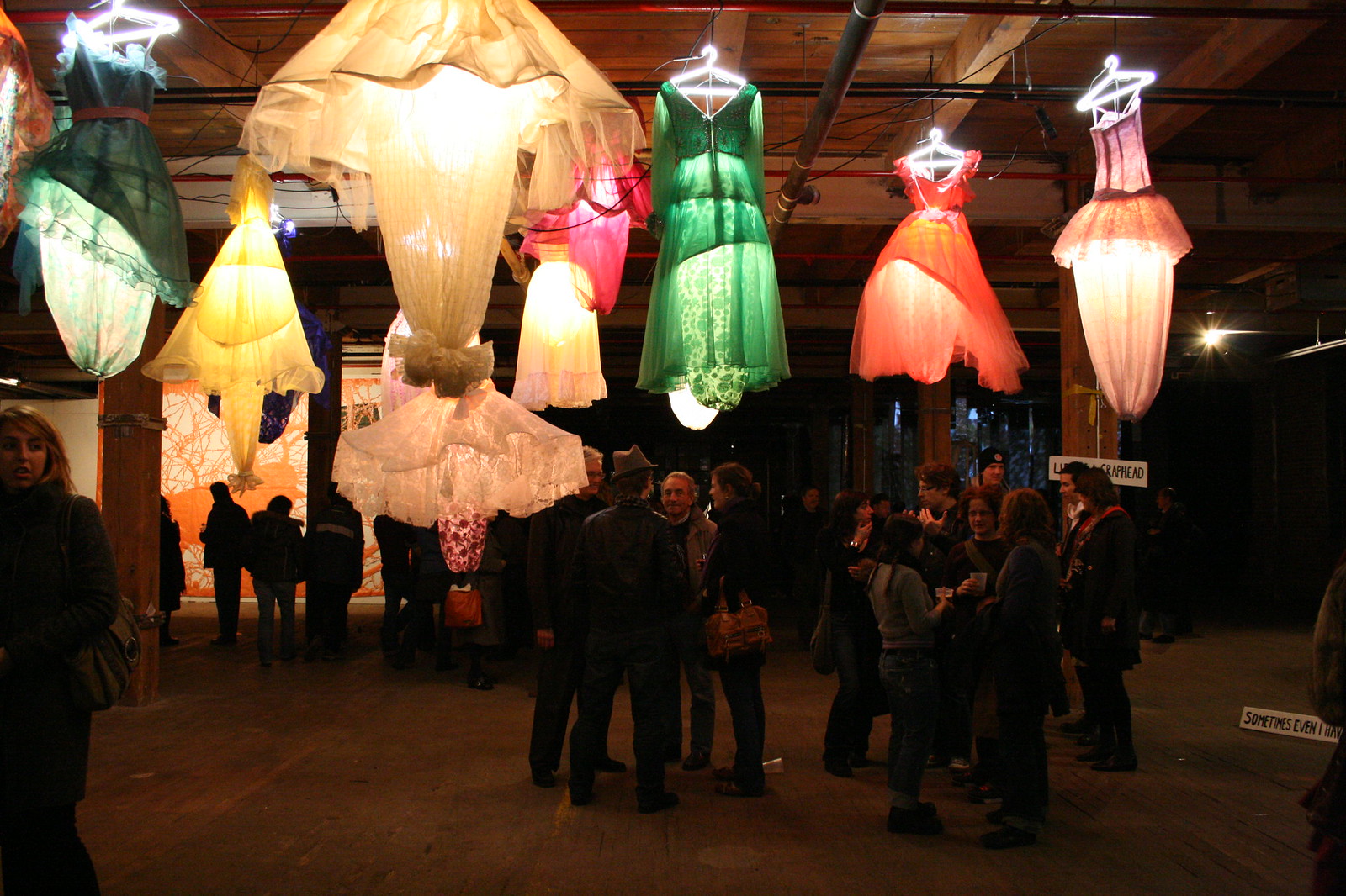 Glowing dresses on hangers suspended as lanterns over a crowd in a dim gallery space.