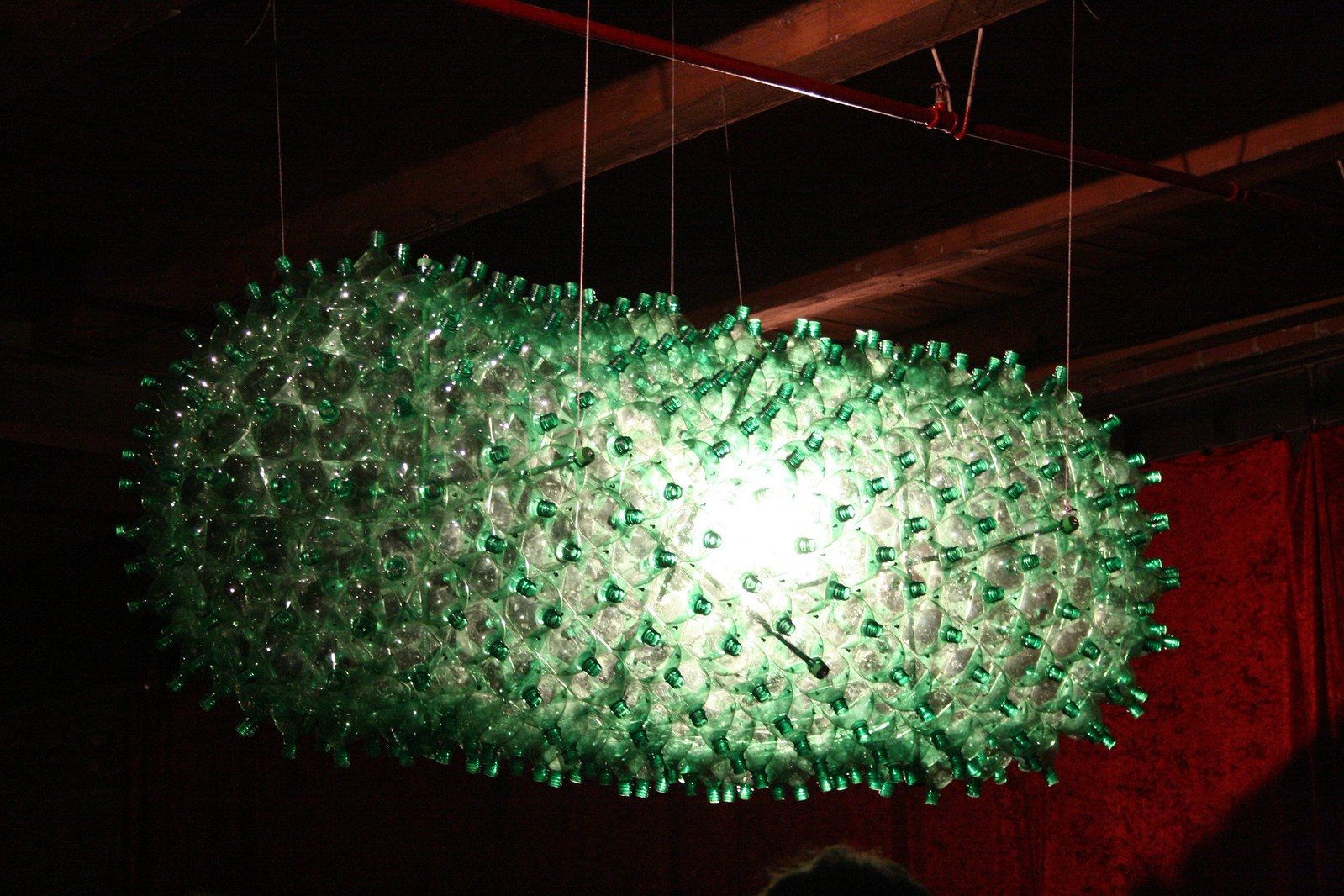 Suspended sculpture made from hundreds of green-capped plastic bottles, glowing from an interior light in a dark room.