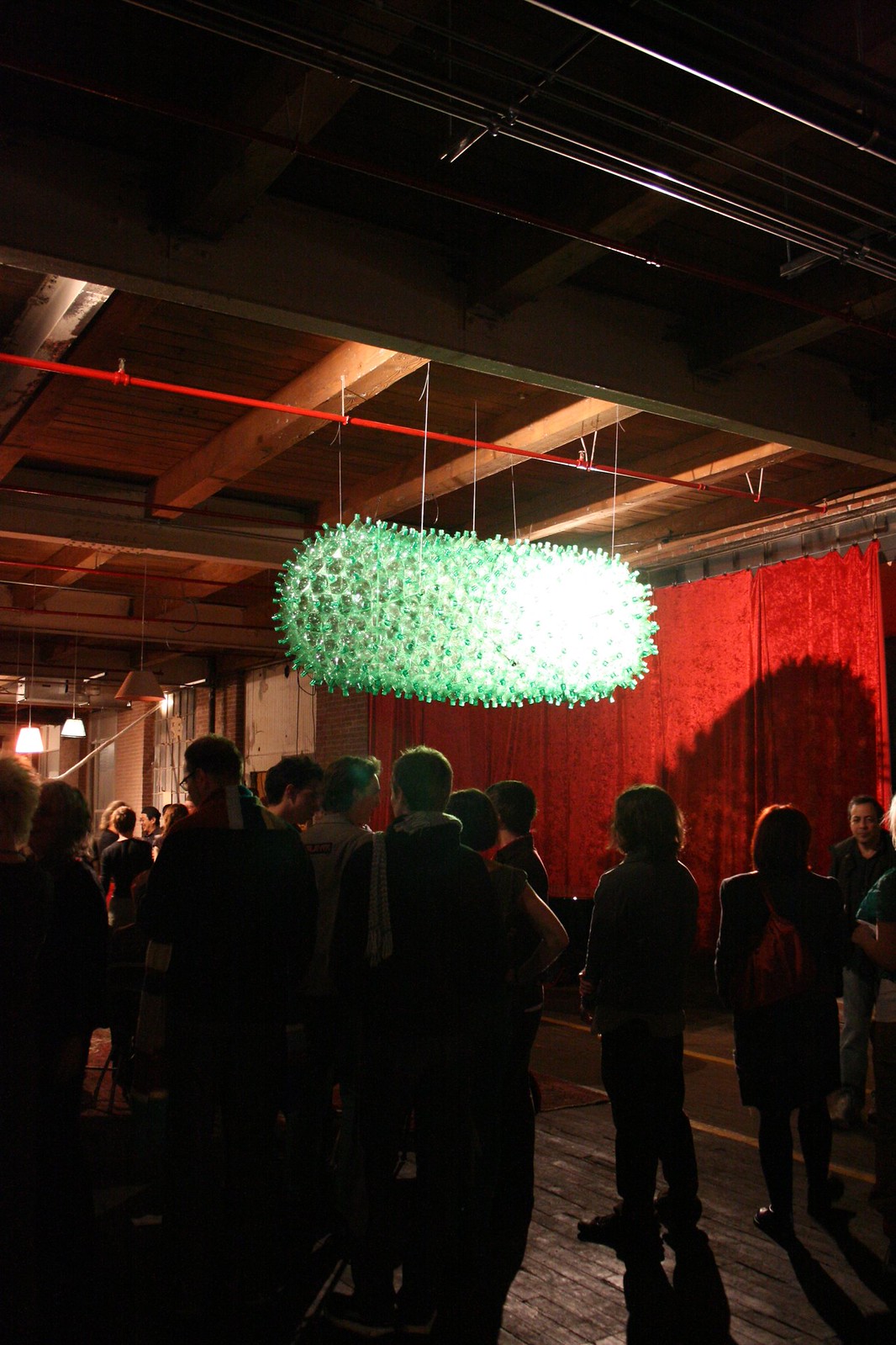 Glowing green suspended sculpture of plastic bottles above a crowd in a dim warehouse gallery with red curtains.