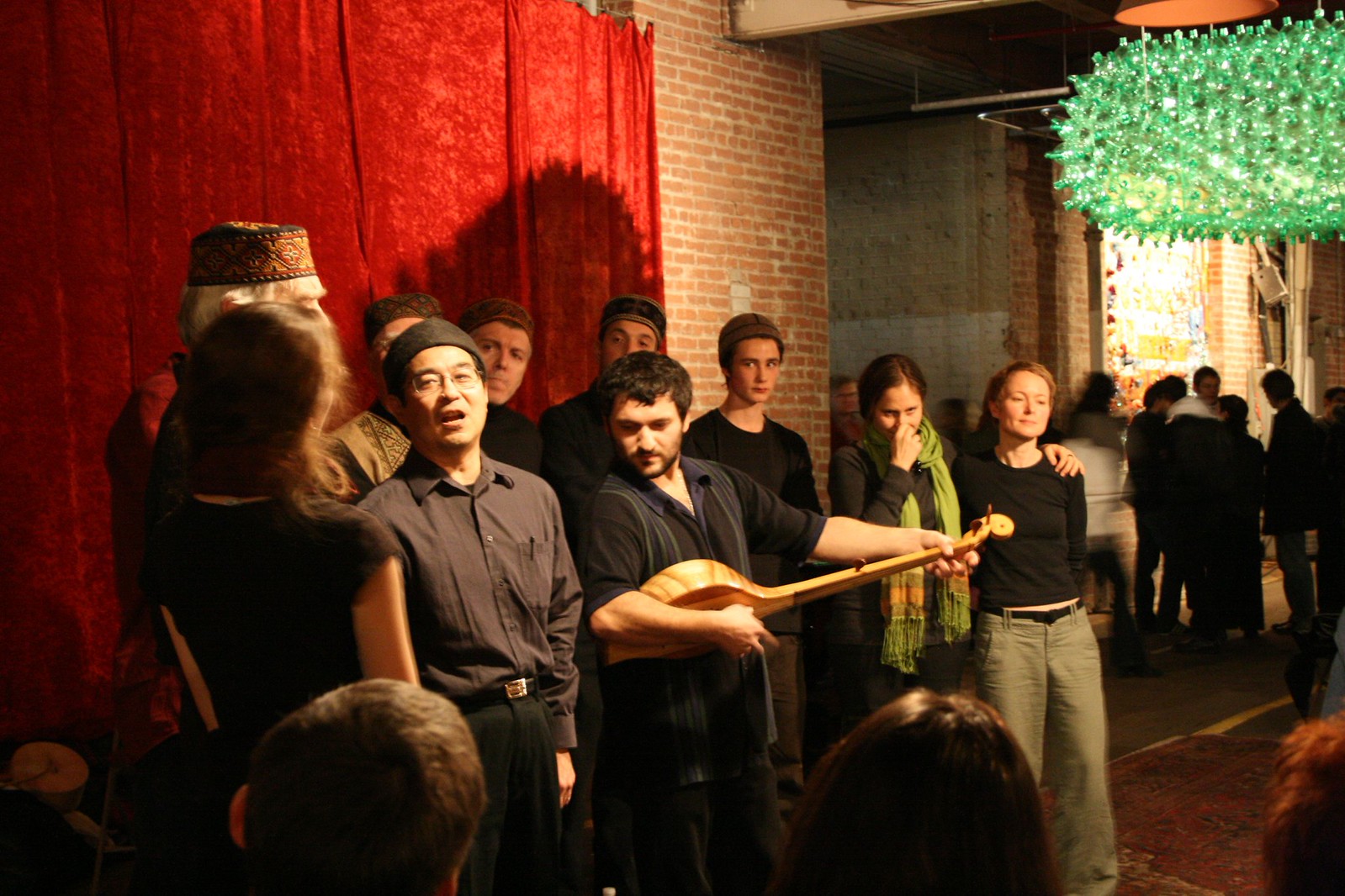A vocal ensemble sings before a red curtain and brick wall as a man holds a long-necked lute.
