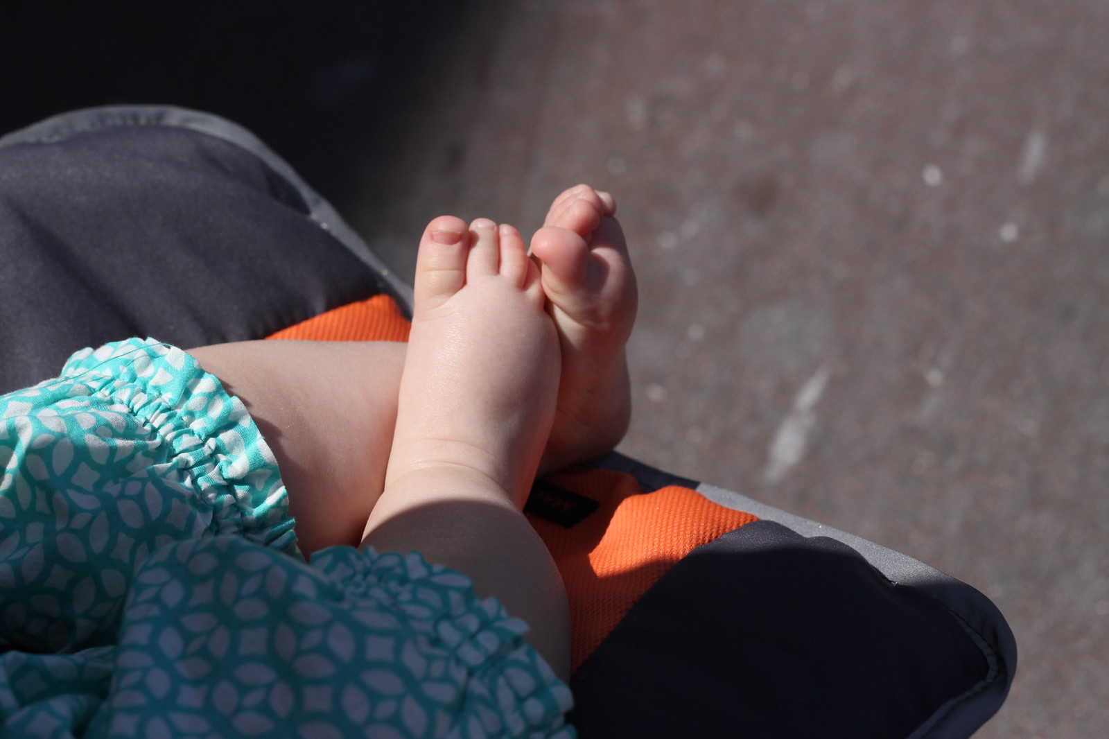 A baby's bare feet crossed at the ankles resting on an orange and gray stroller, in teal-patterned clothes.