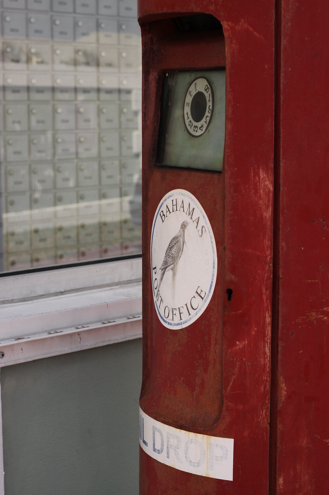 Weathered red mailbox with a "Bahamas Post Office" bird decal and a "MAIL DROP" sticker beside a wall of PO boxes.