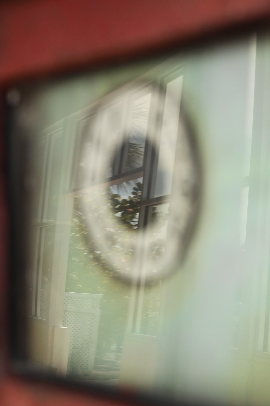 A trees-and-sky reflection glimpsed through a paned window and a wreath, framed by a red wall.