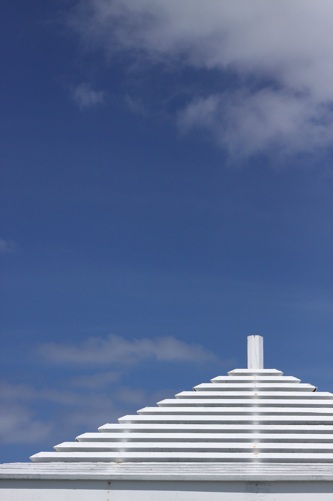 White stepped pyramid-shaped wooden roof with a small finial against a bright blue sky with wispy clouds.