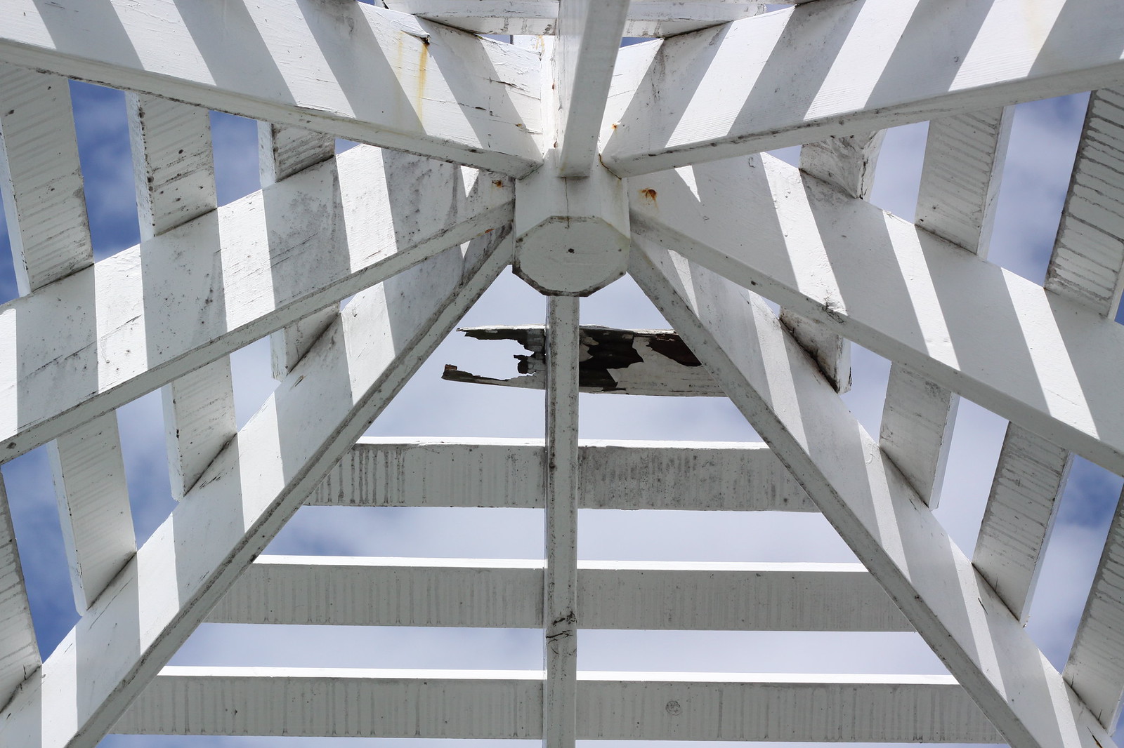 Looking up at the underside of a white wooden gazebo roof against blue sky, with peeling paint and a broken slat.