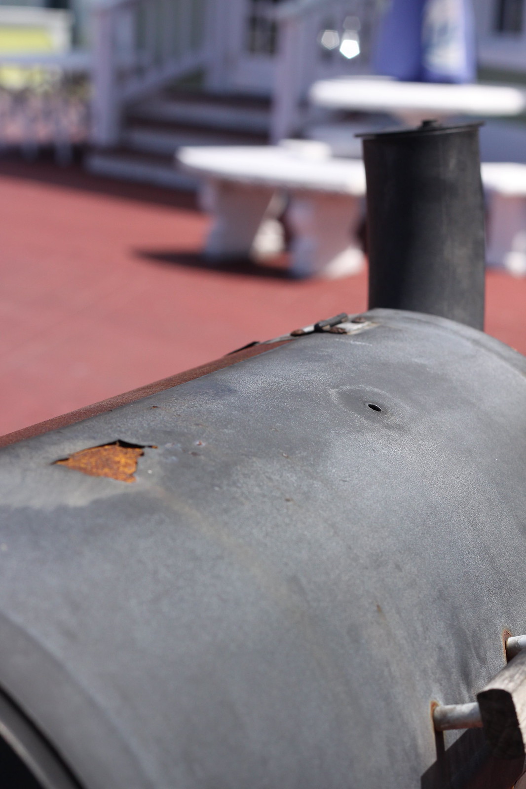 Close-up of a weathered metal barrel smoker with rust spots and a chimney on a patio with benches.