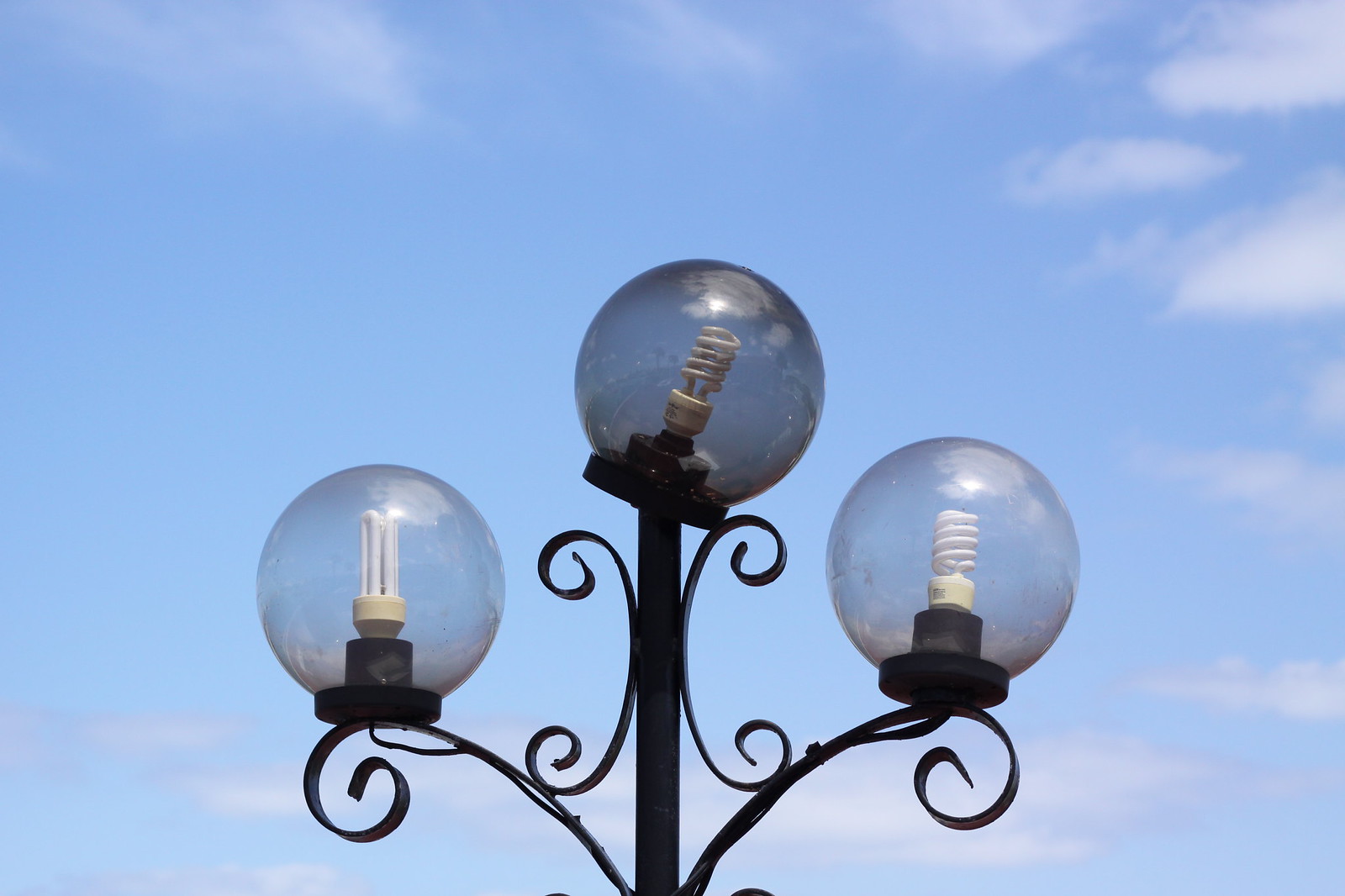 Ornate wrought-iron lamppost with three glass globe lights and CFL bulbs against a blue sky.