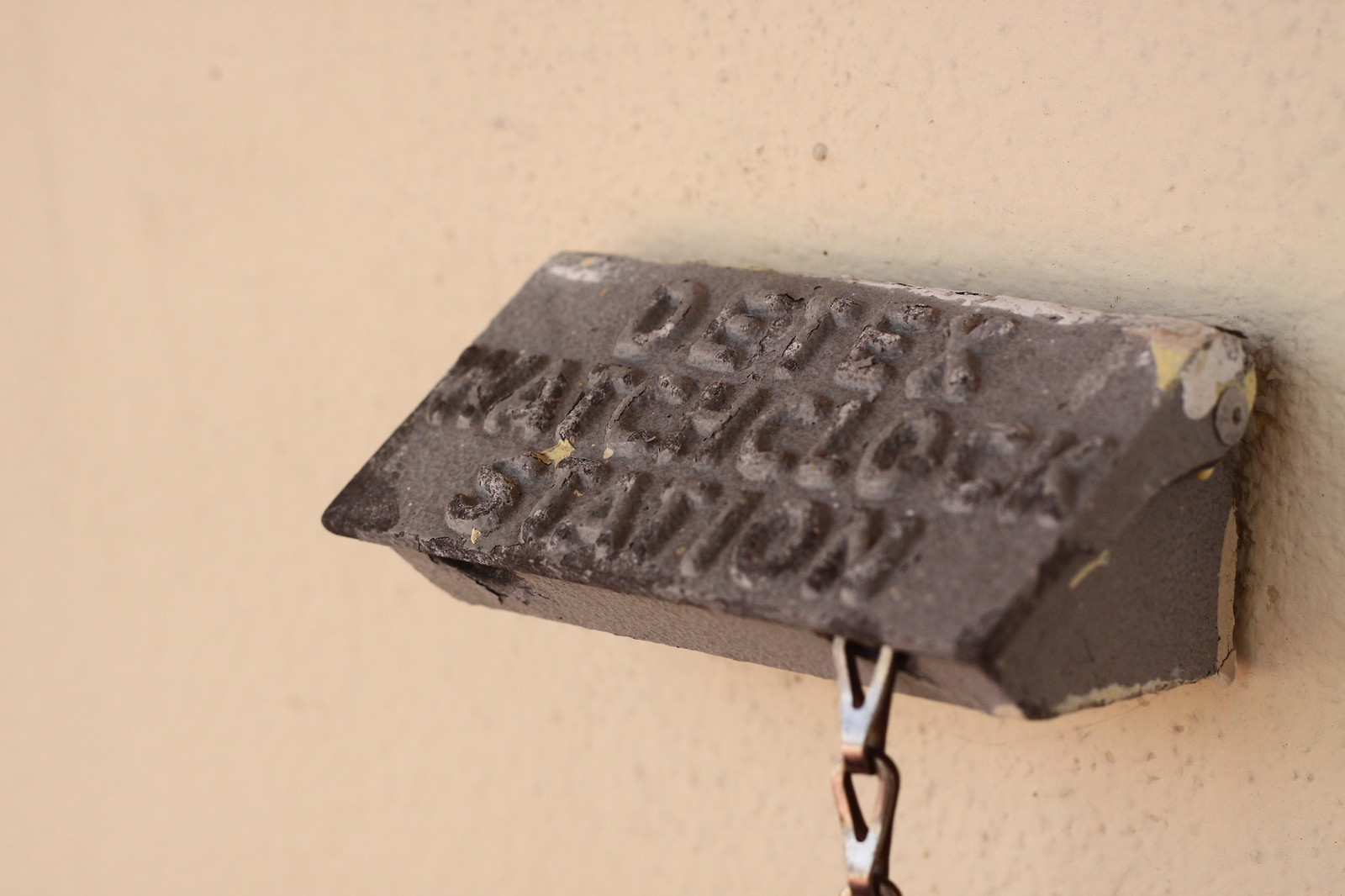 Weathered cast-iron "Detex Watchclock Station" plaque mounted on a beige wall with a hanging chain below.