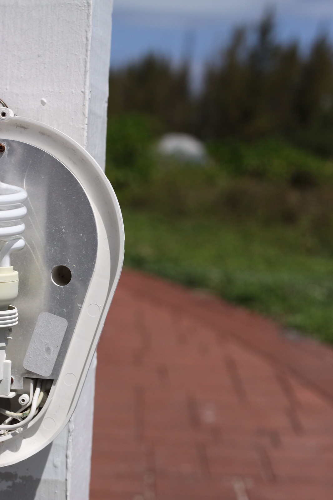 A coverless outdoor light fixture with an exposed bulb on a white wall, overlooking a brick path and greenery.