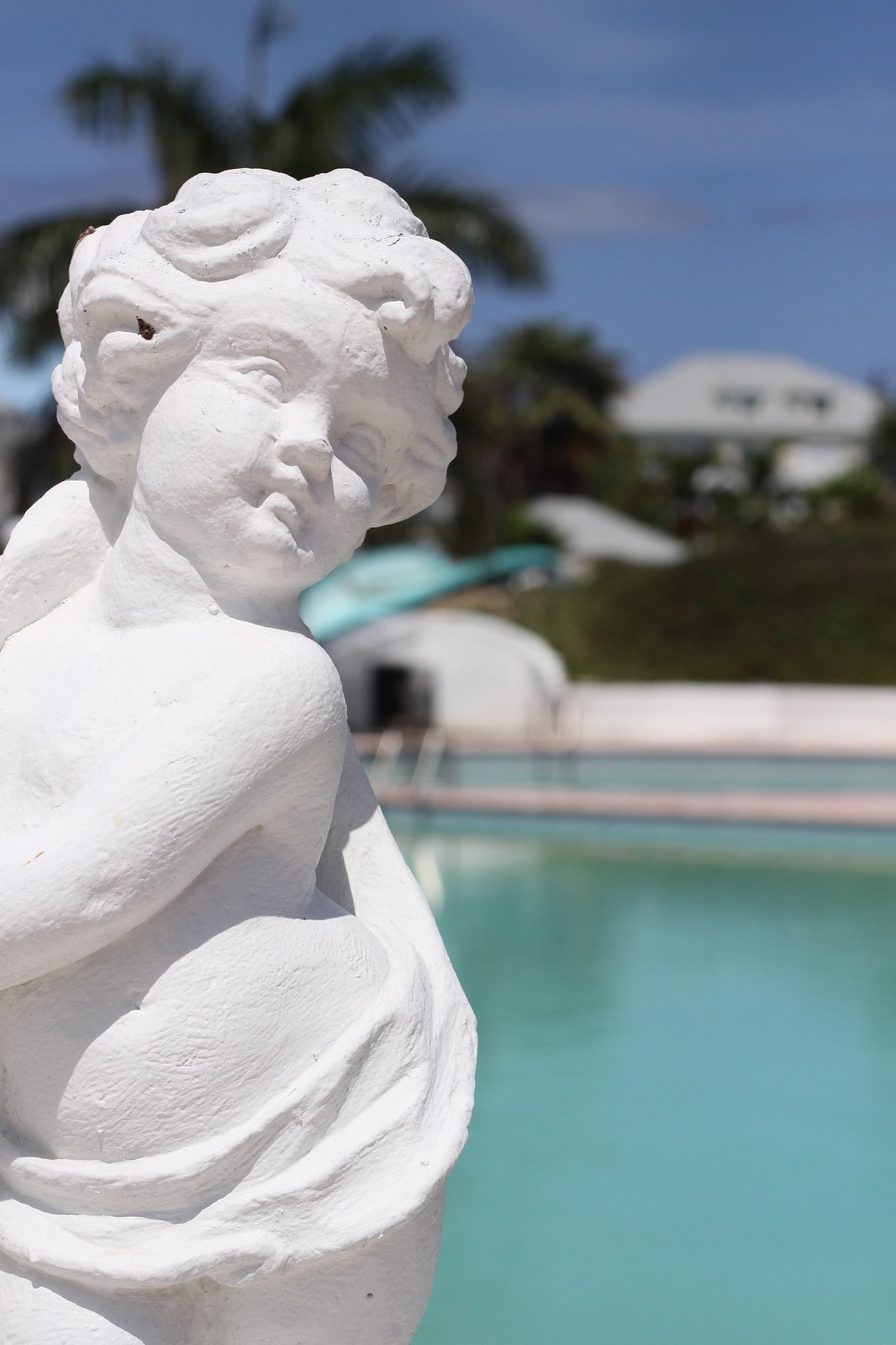 White cherub statue beside a turquoise pool, with palm trees and houses under a blue sky.
