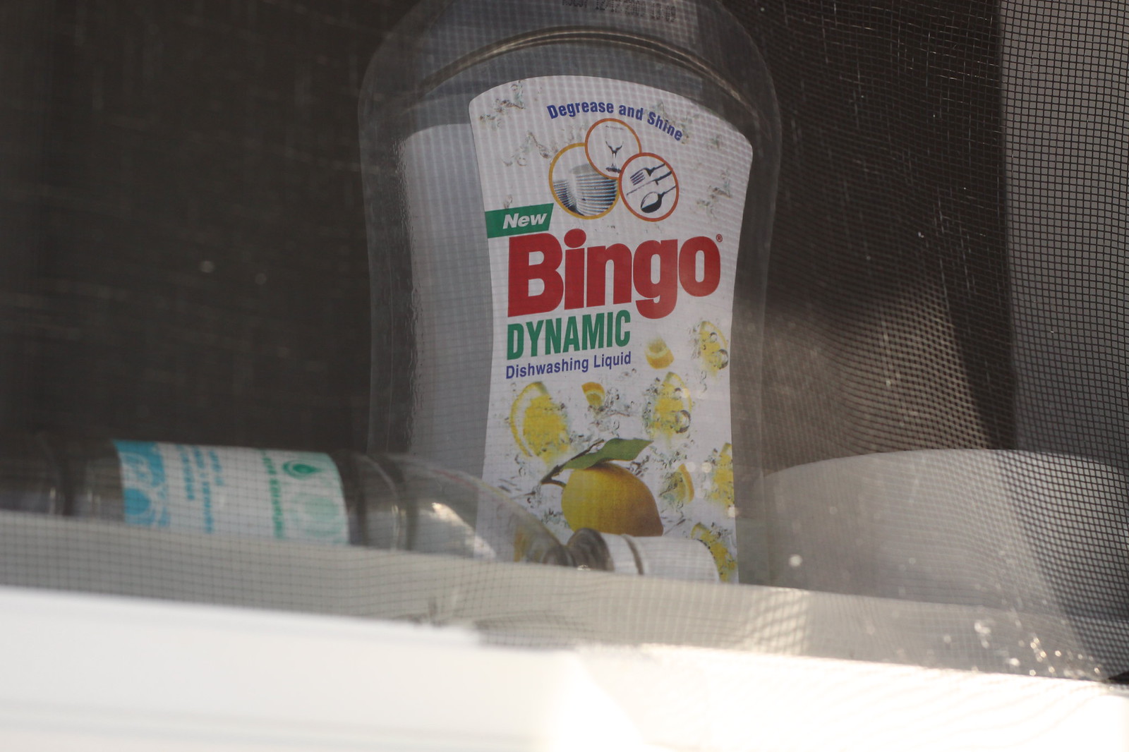 A bottle of "Bingo Dynamic" lemon dishwashing liquid on a windowsill behind a screen.