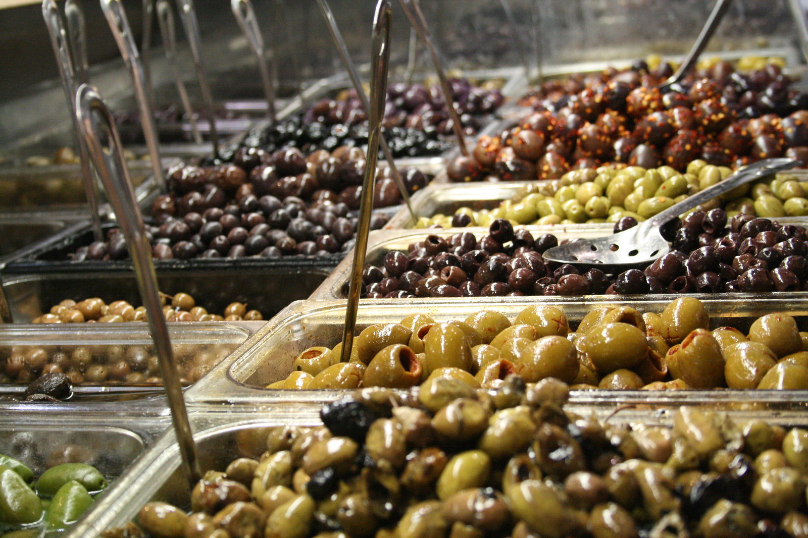 Trays of green, black, and marinated olives with metal tongs and slotted spoons at a market deli counter.